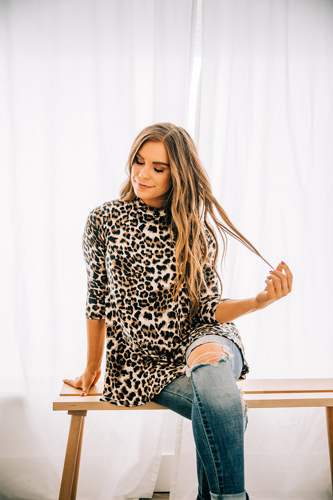  cheetah print peplum top online clothing boutique commercial photography station studio logan utah model with long beachy waves holding strands of hair crossing legs distressed jeans looking down sitting on a bench commercial clothing boutique #onli