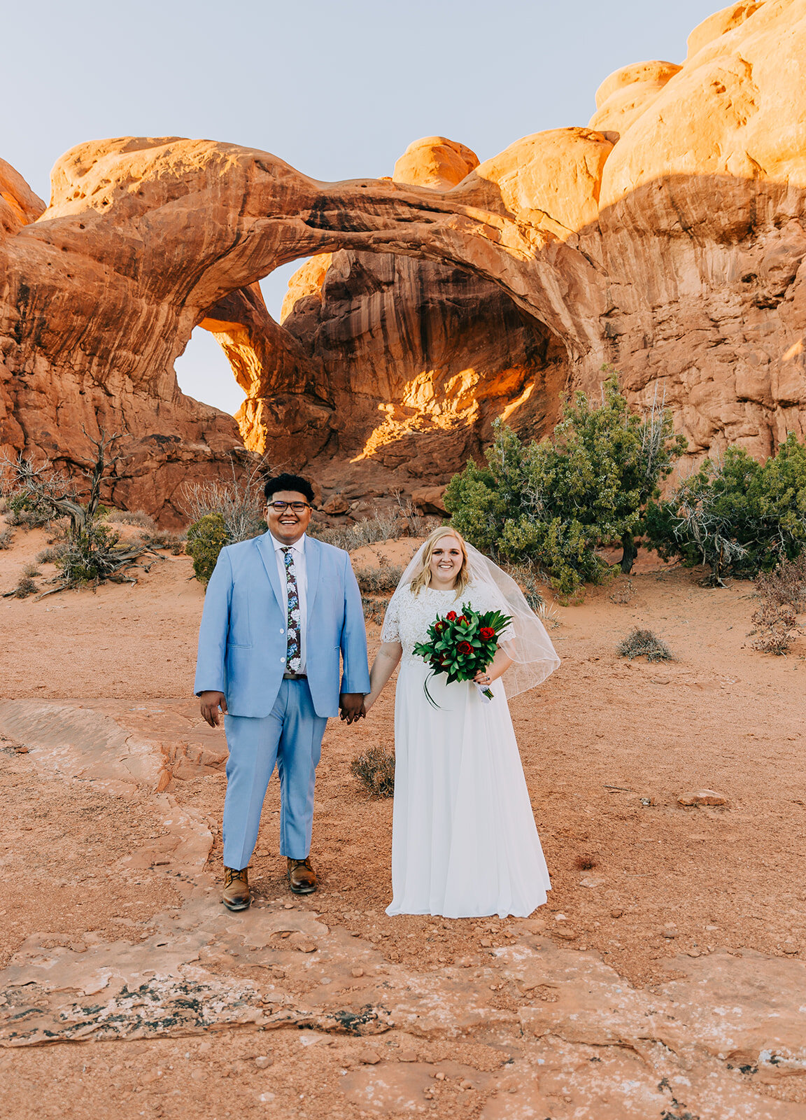  bride and groom standing and holding hands pose inspiration desert moab utah powder blue suit groom fashion bridal fashion modest wedding dress white dress veil bouquet inspiration double arch elephant rock professional photographers in utah utah we