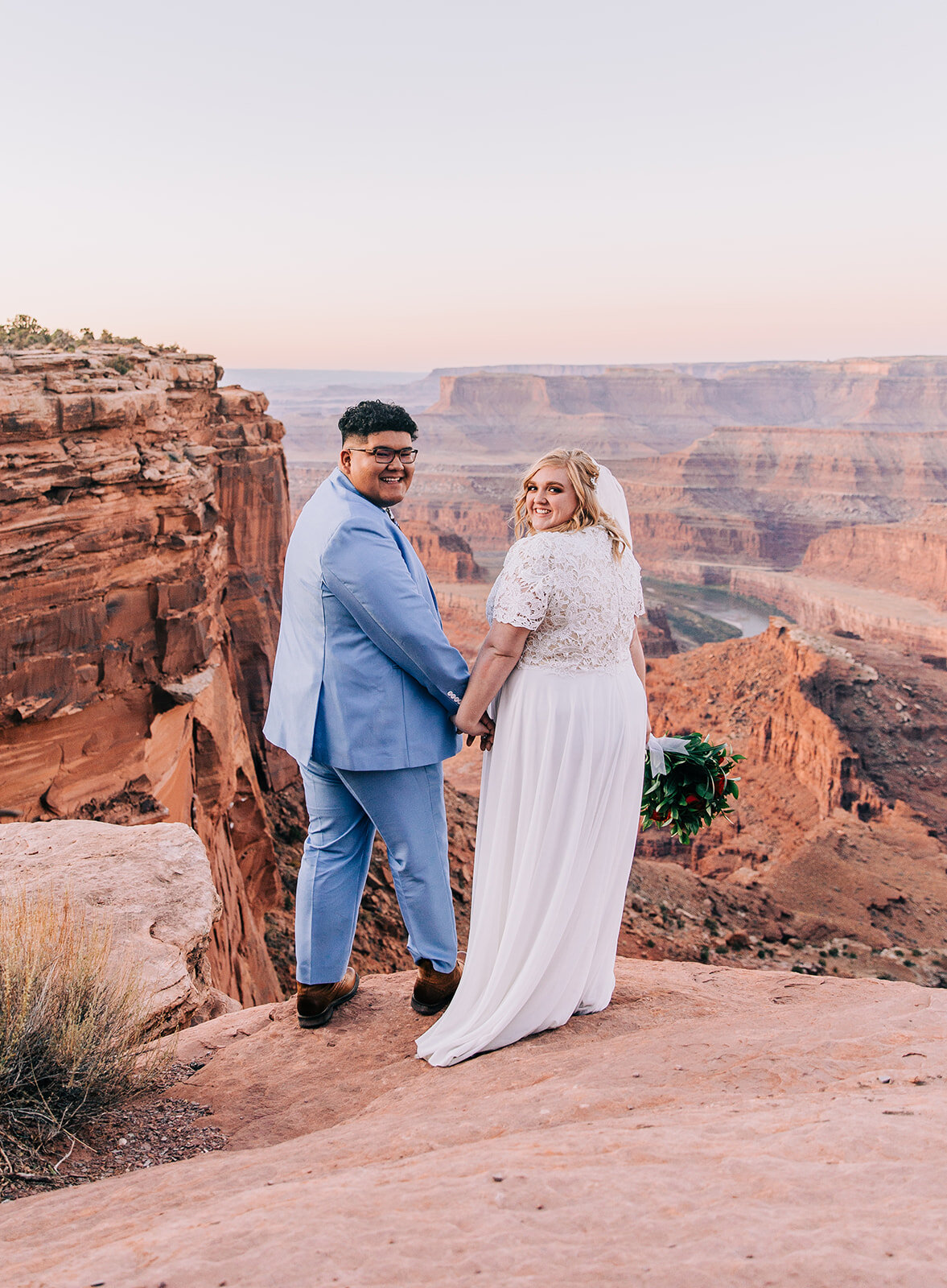  couple standing on a ledge over the shoulder smiling pose inspo moab utah groom smiling powder blue suit groom fashion modest wedding dress wedding dress inspo white dress veil bouquet inspo double arch elephant rock professional photographers in ut