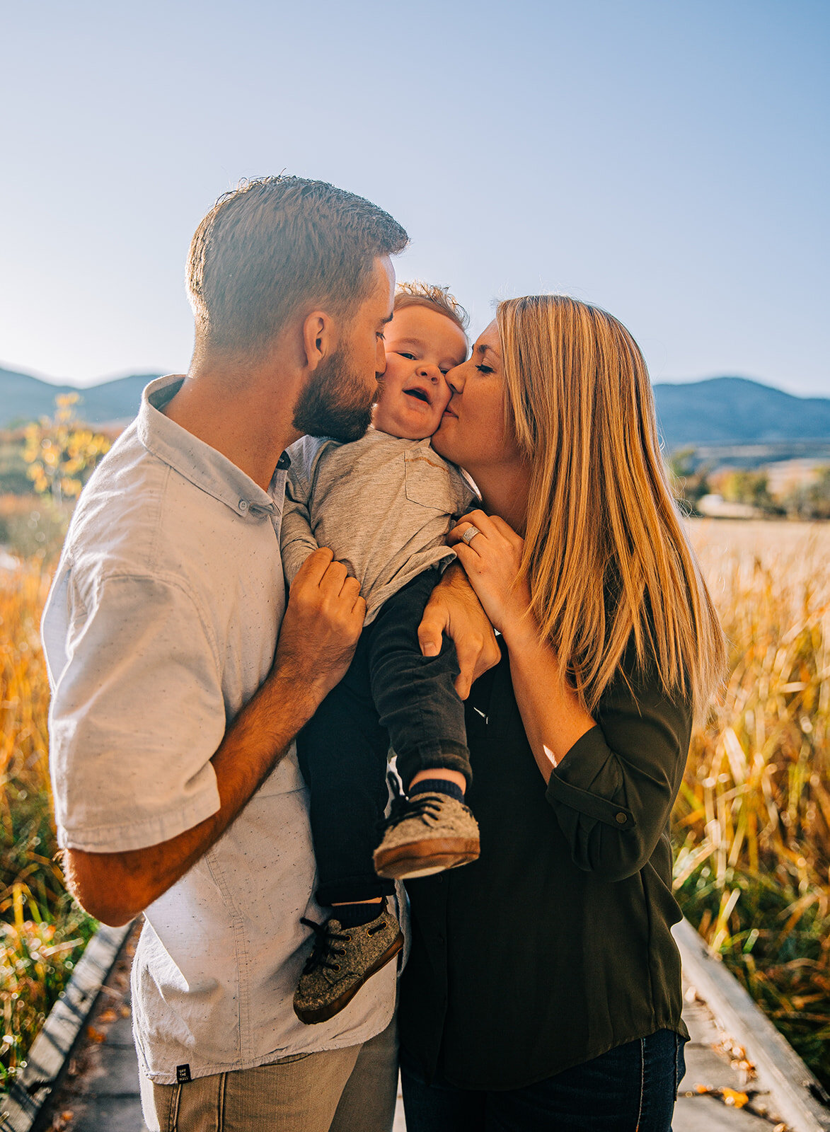  family of three boardwalk family pictures garden city park bear lake utah together again mom and dad holding baby and kissing cheeks fields of gold mountains lakeside photos professional photographers in utah utah family photographers extended famil