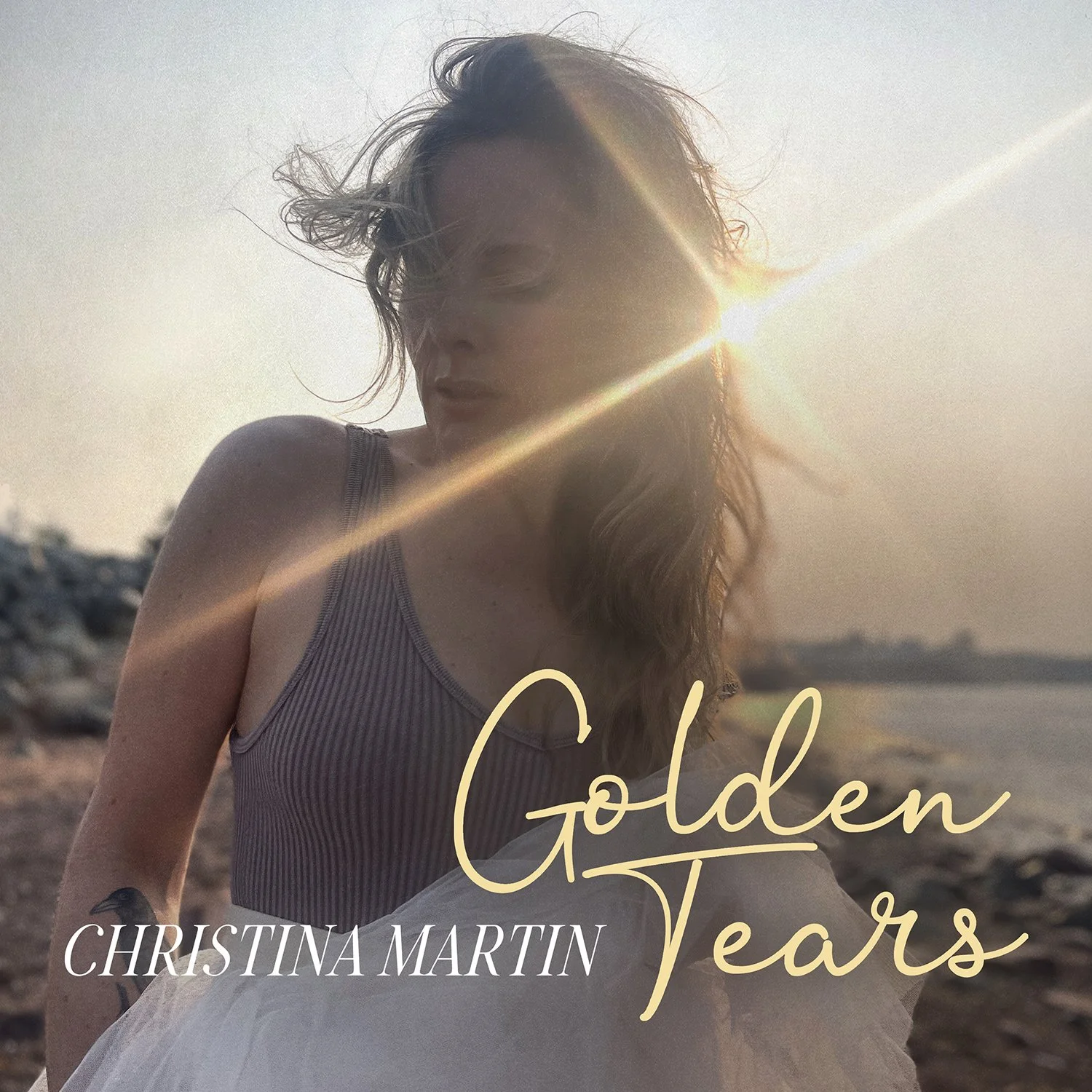Woman standing by the ocean at sunset with wind in her hair and sun rays across her face, wearing a sleeveless top and tulle skirt, with the text “Golden Tears – Christina Martin.”
