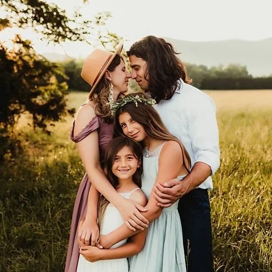 A family of four in a field, with a woman and man embracing and two young girls smiling in front.