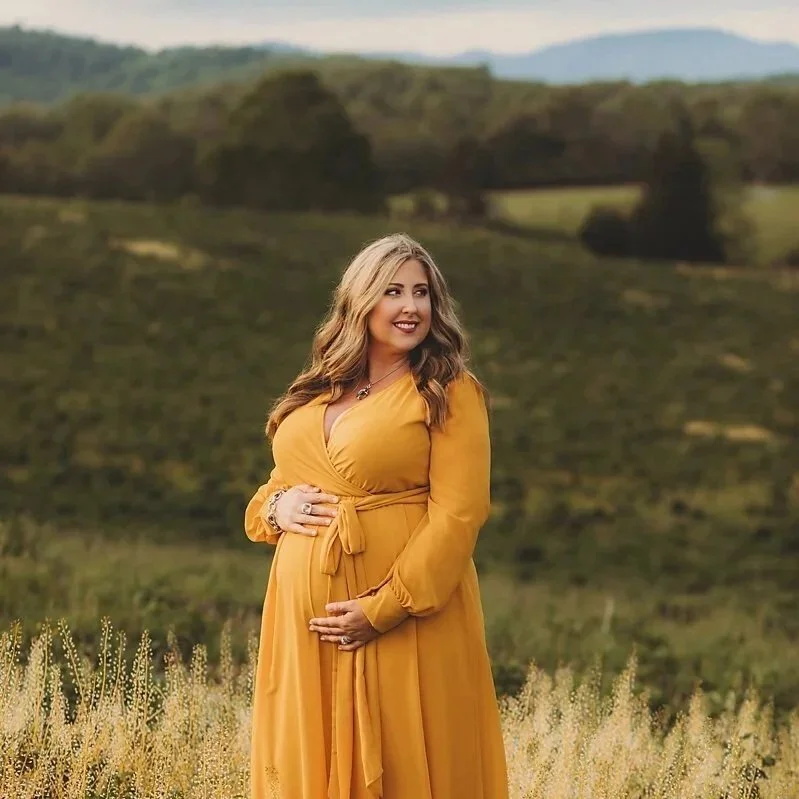 A pregnant woman in a mustard yellow dress standing outdoors in a field with green hills in the background, smiling and looking to her right.