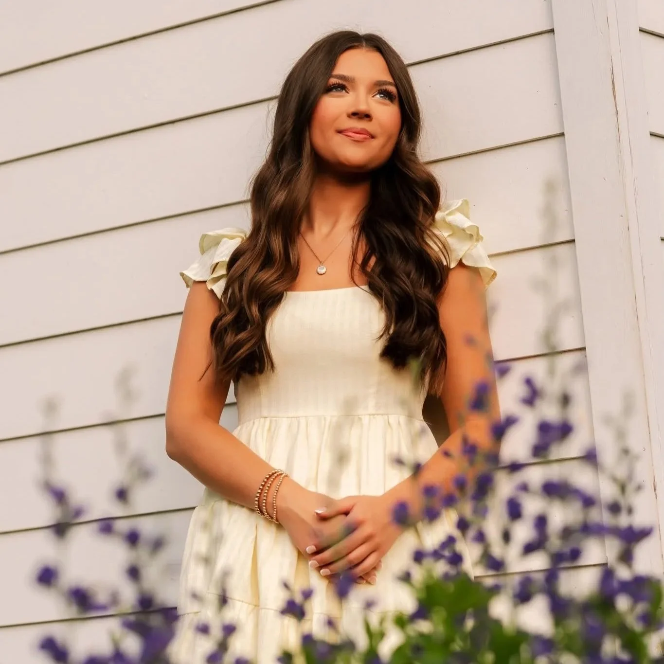 A young woman with long wavy brown hair is standing outdoors in front of a white wooden siding wall, wearing a light yellow dress with ruffled sleeves, jewelry including a pendant necklace, bracelets, and rings, and she is smiling softly with her hands clasped in front of her.