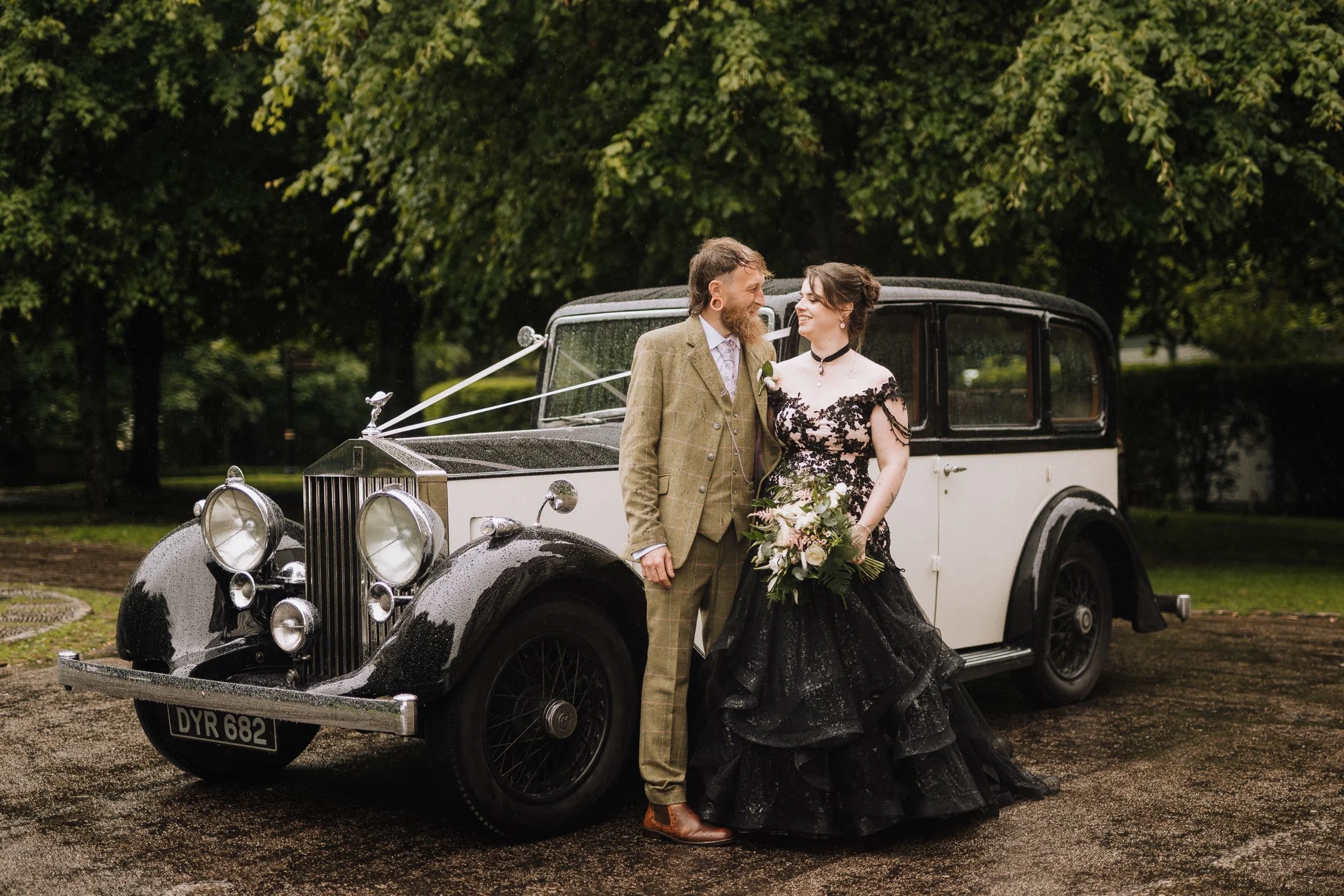 A couple dressed in vintage wedding attire standing next to a classic black and white car outdoors, with green trees in the background. The woman holds a bouquet, and both are smiling at each other.
