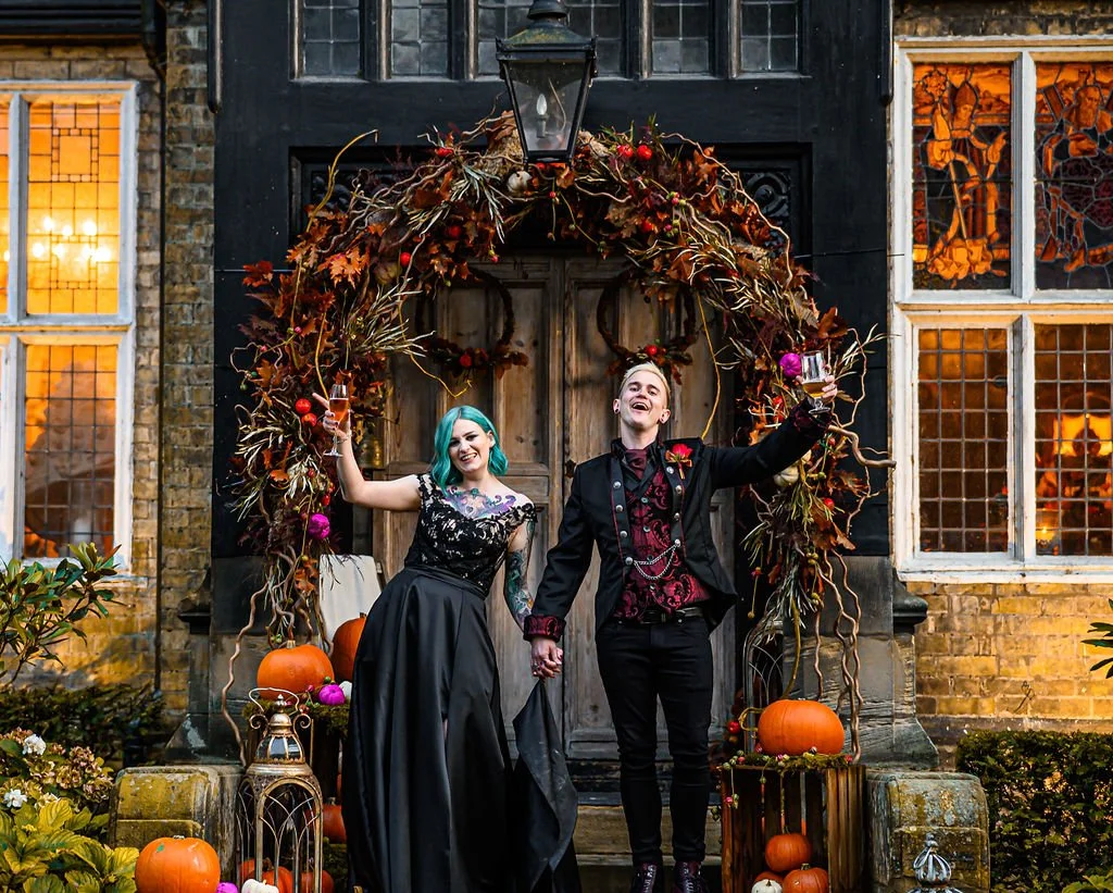 A couple dressed in gothic attire celebrating outdoors at night, holding hands and raising glasses in front of a decorated arch with pumpkins and vines.