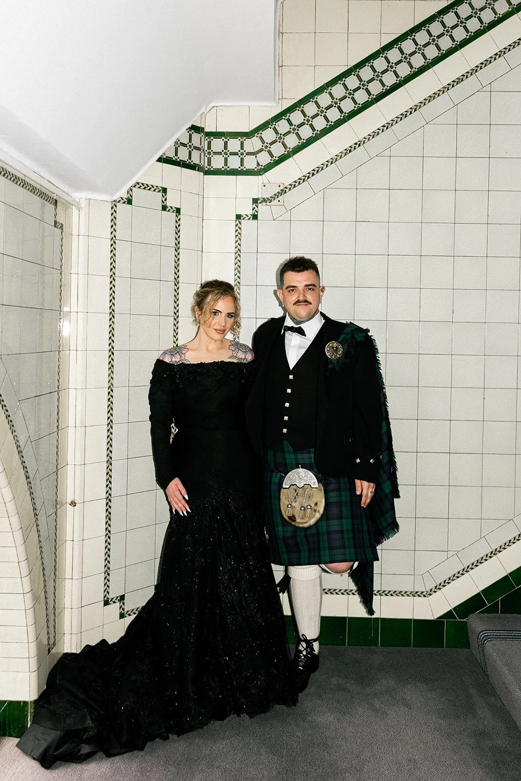 couple stand at the bottom of a stairwell leaning against the tile wall. Bride wears black glittery mermaid gown while the groom wears formal highland wedding outfit in blackwatch tartan