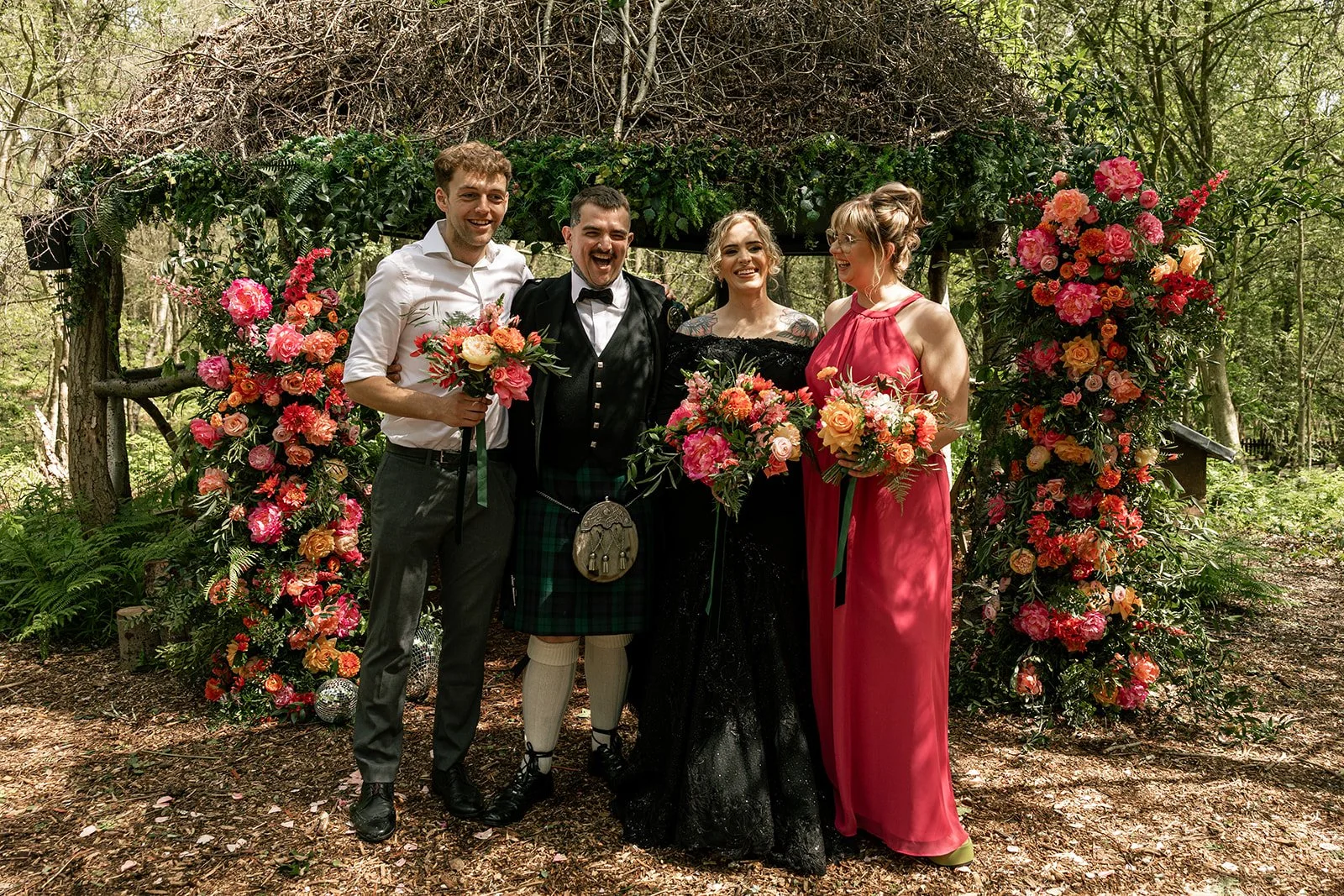 Couple stand with bridesmaid and bestman in front of the woodland wedding arch decorated with pink and orange wedding flowers and glitter balls, everyone except the groom carries a bouquet