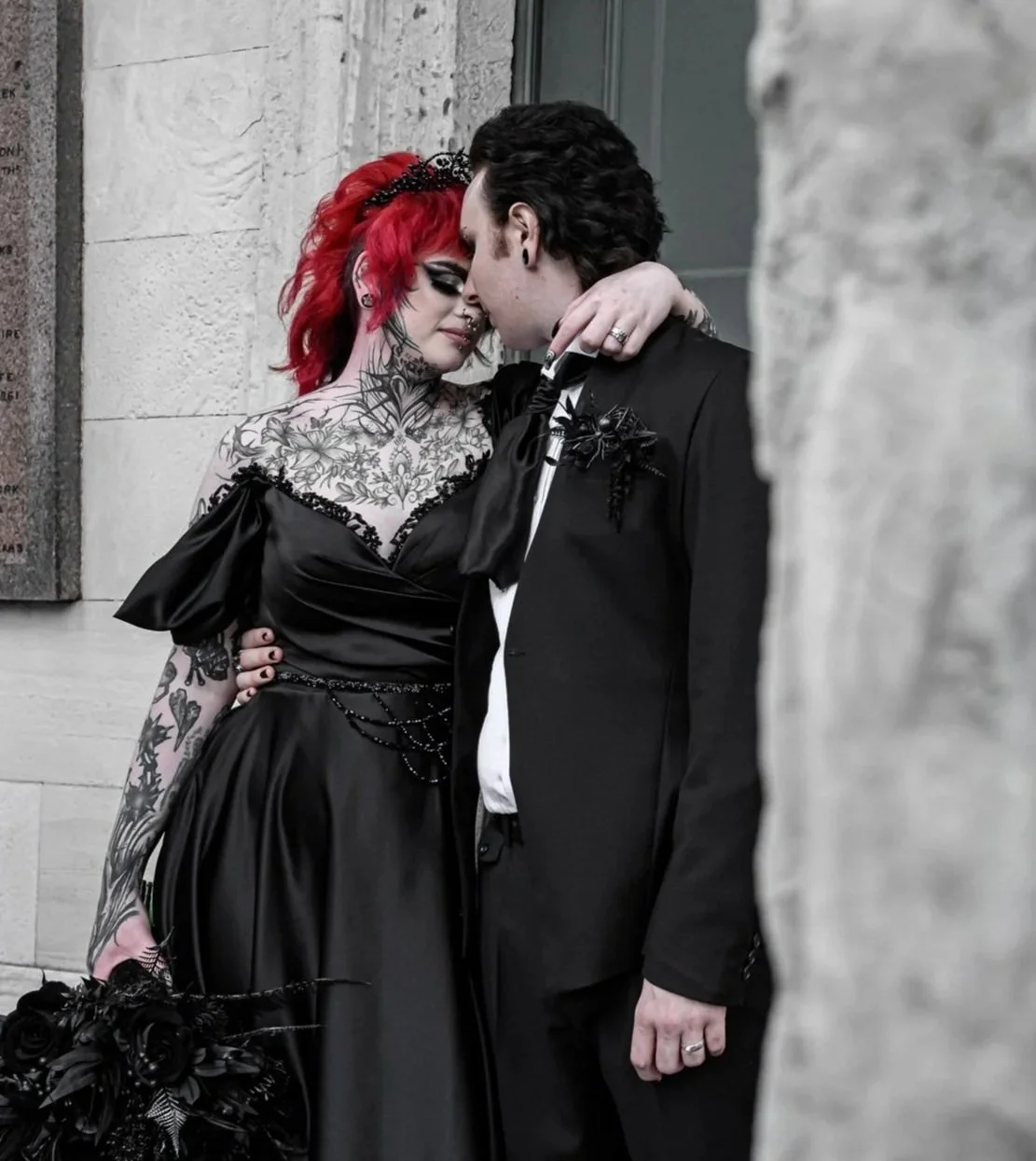 A couple dressed in gothic style wedding clothing, closely embracing and about to kiss against a stone wall.