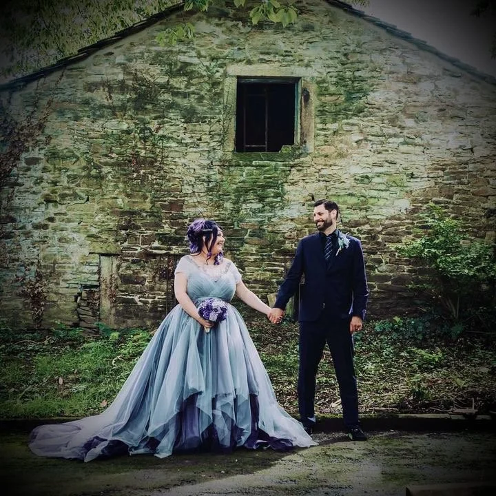 A bride and groom holding hands and smiling at each other outdoors in front of an old stone building with greenery.