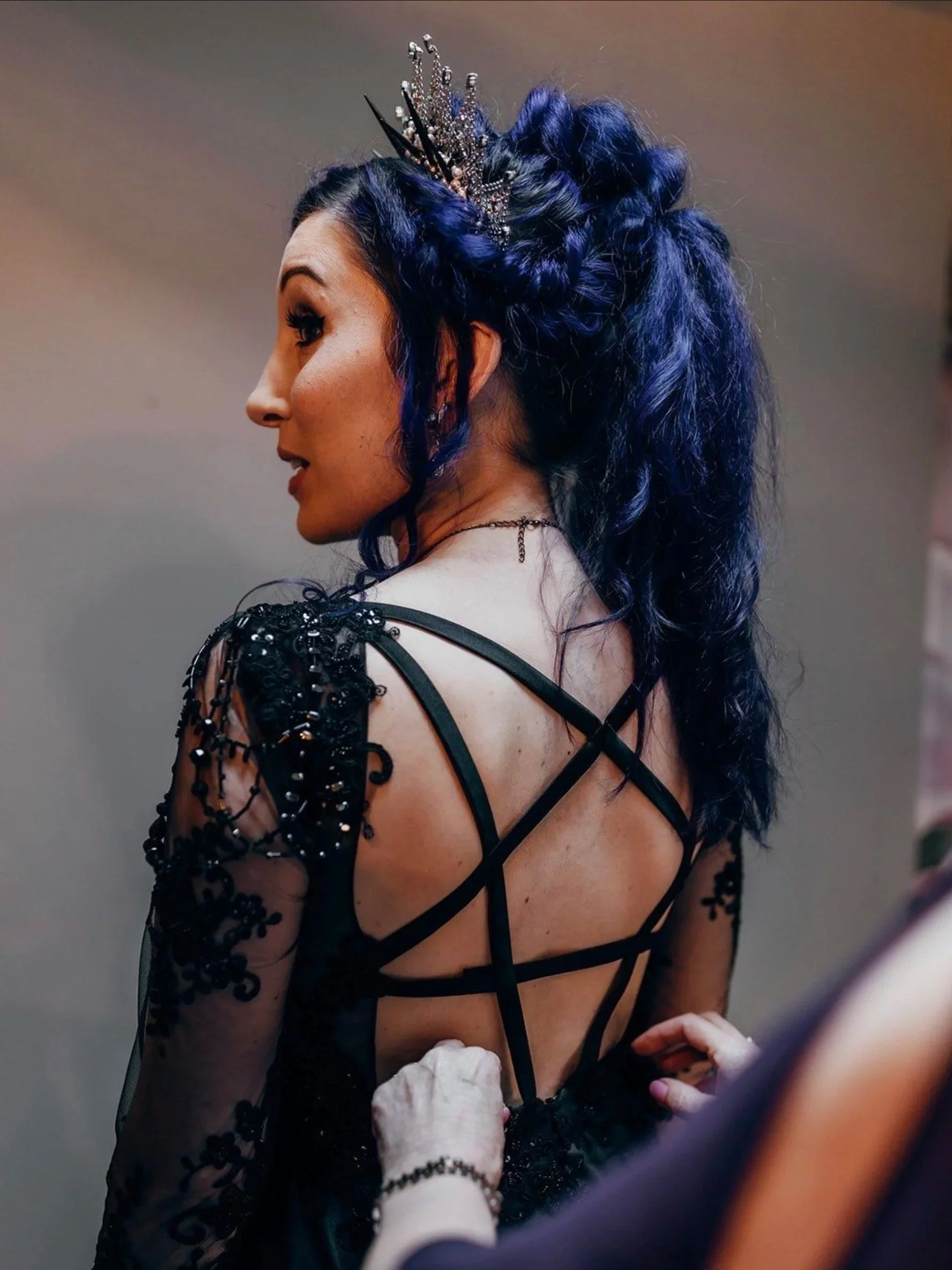 A woman with dark blue, curly hair styled in an updo, wearing a sparkly tiara and a black, sheer dress with lace and beaded embellishments, being assisted with her dress by a person with a white glove.