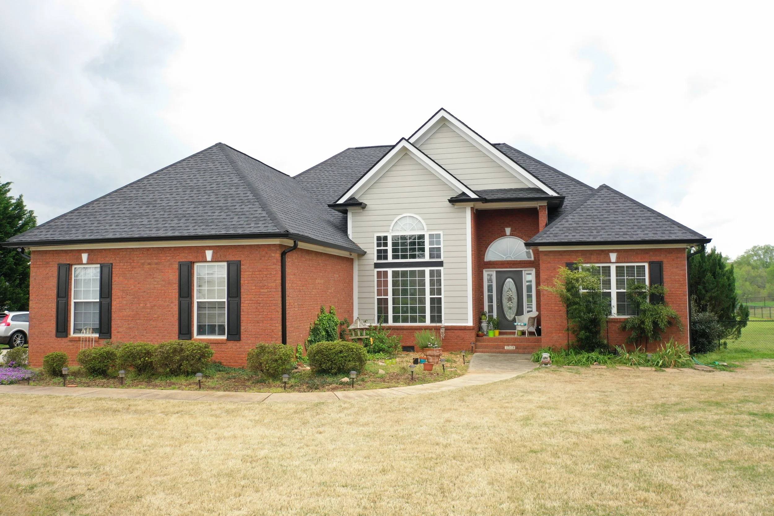 A two-story house with a brick lower level and a gray siding upper level. The house features several large windows, a front door with glass, and a small porch with a chair and potted plants. The yard has a grass lawn and a garden with small bushes and plants.