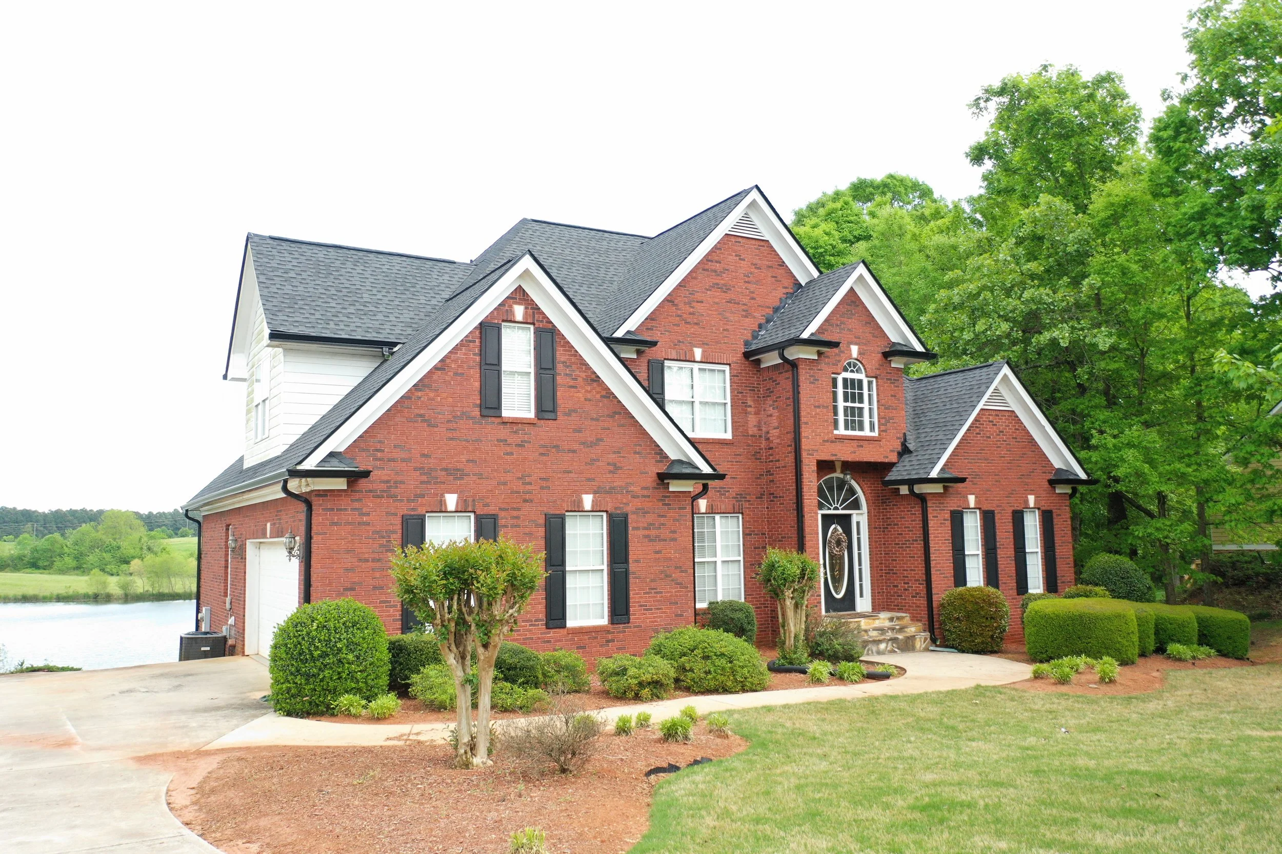 A finished Alumni Roofing and Gutters roofing project. A two-story brick house with black shutters, white trim, and a gray shingled roof, surrounded by green trees and a landscaped yard.