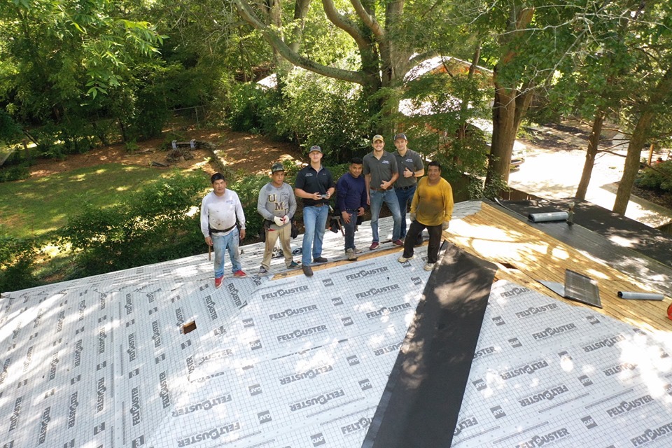 The Alumni Roofing Team standing on a roof under construction, with trees and a backyard visible in the background.