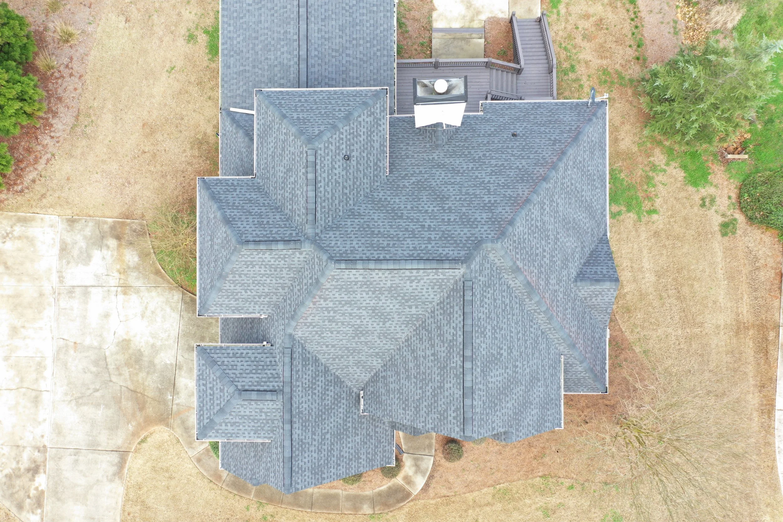 An aerial view of a house with a grey shingle roof, surrounded by patches of grass, dirt, and trees, with part of a concrete driveway visible in the lower left corner.