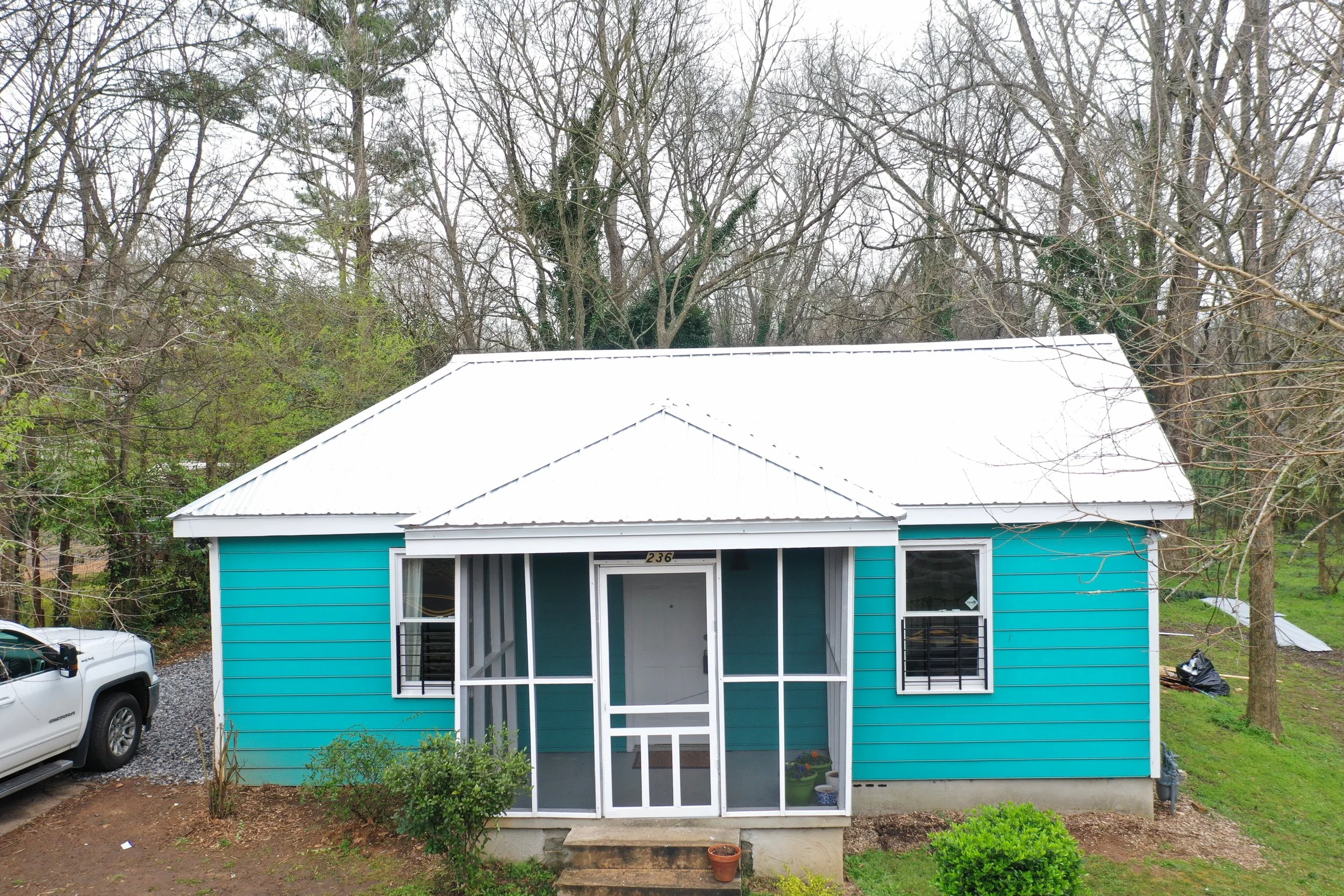 A small house with bright turquoise siding and white trim, featuring a screen door, situated in a yard with starting to bloom shrubs and a concrete step leading up to the porch. The house number "236" is displayed above the door. The background shows