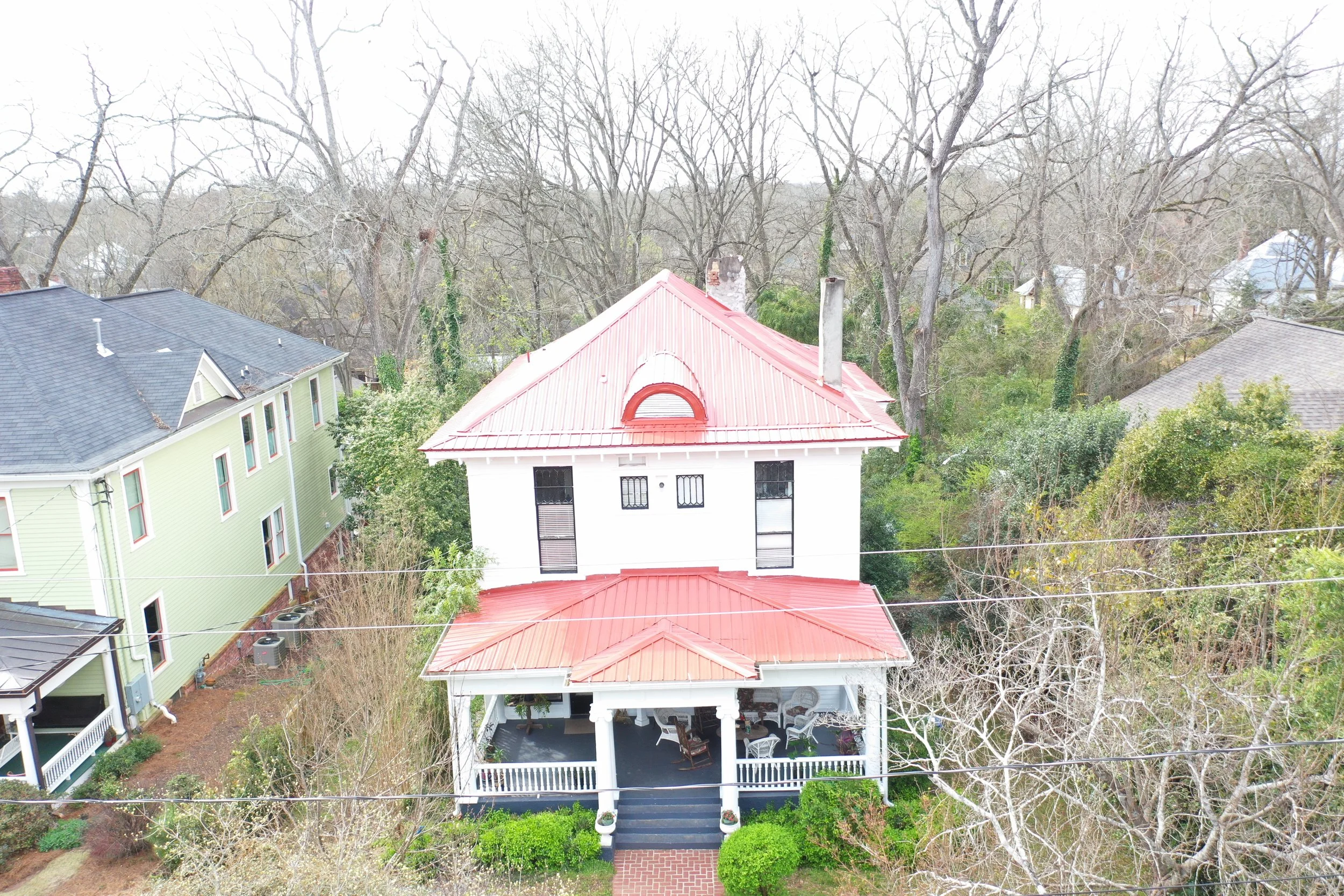 Aerial view of a white house with a red roof, porch, and front steps, surrounded by trees and neighboring houses in a residential neighborhood.