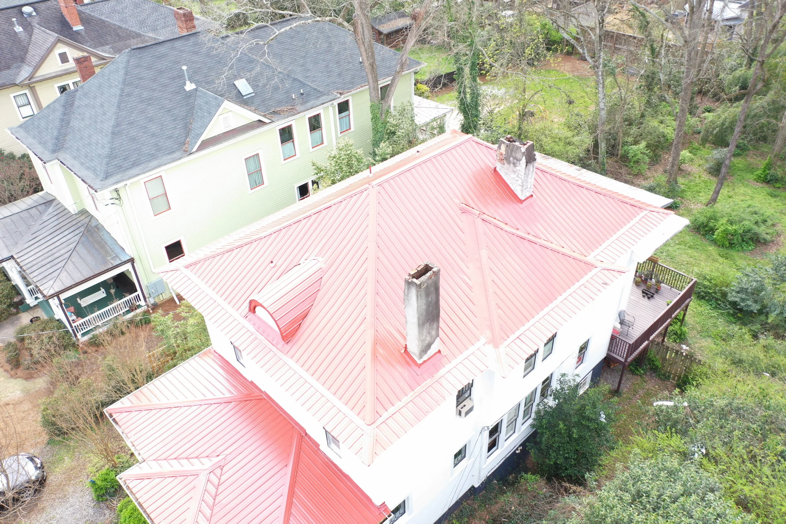 Aerial view of a house with a red metal roof surrounded by trees and other houses with gray roofs.