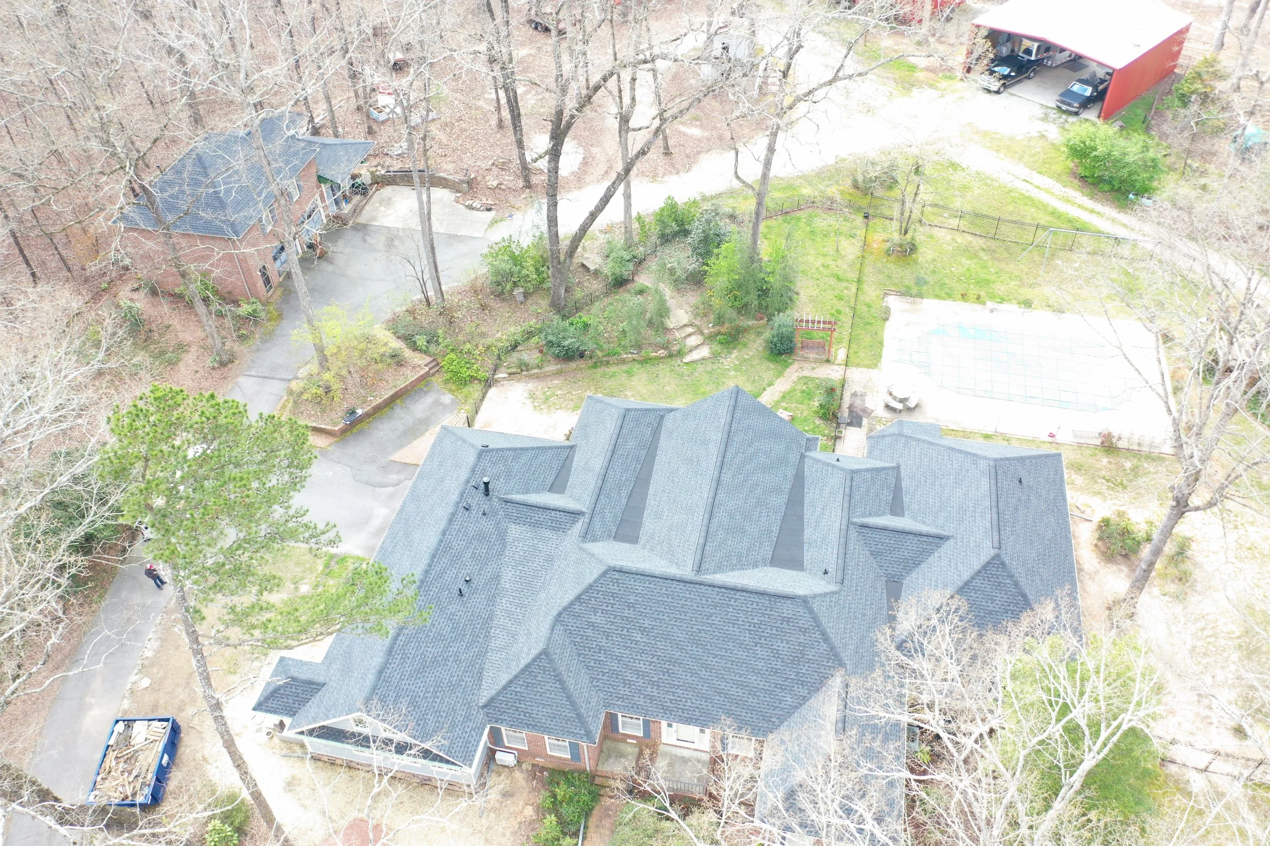 Aerial view of residential neighborhood showing trees, houses with gray roofs, a backyard with a tennis court, and a large red shed with cars parked inside.