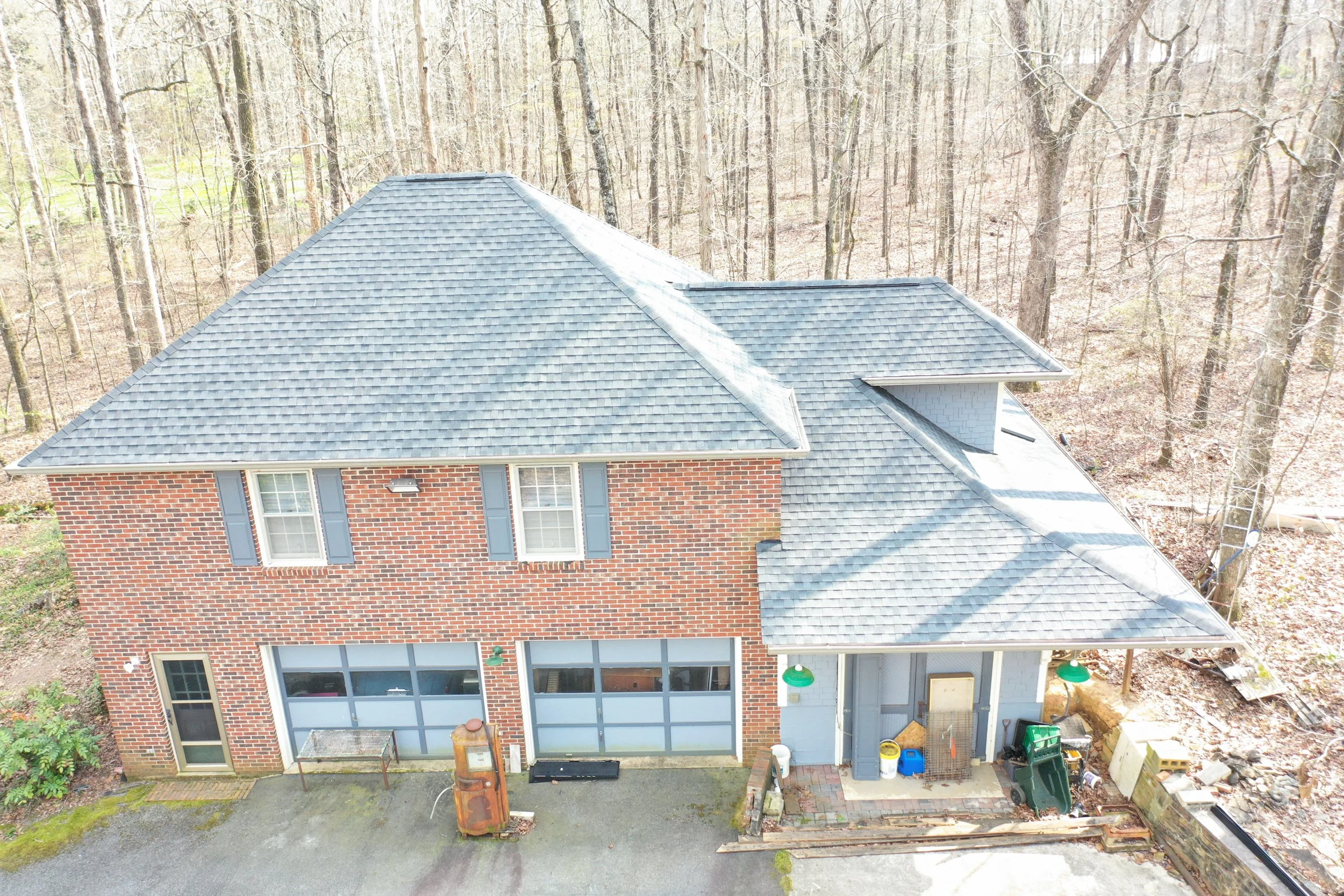 Aerial view of a two-story brick house with a gray shingled roof, surrounded by a wooded area with leafless trees, a small patio with outdoor furniture, and various yard items.
