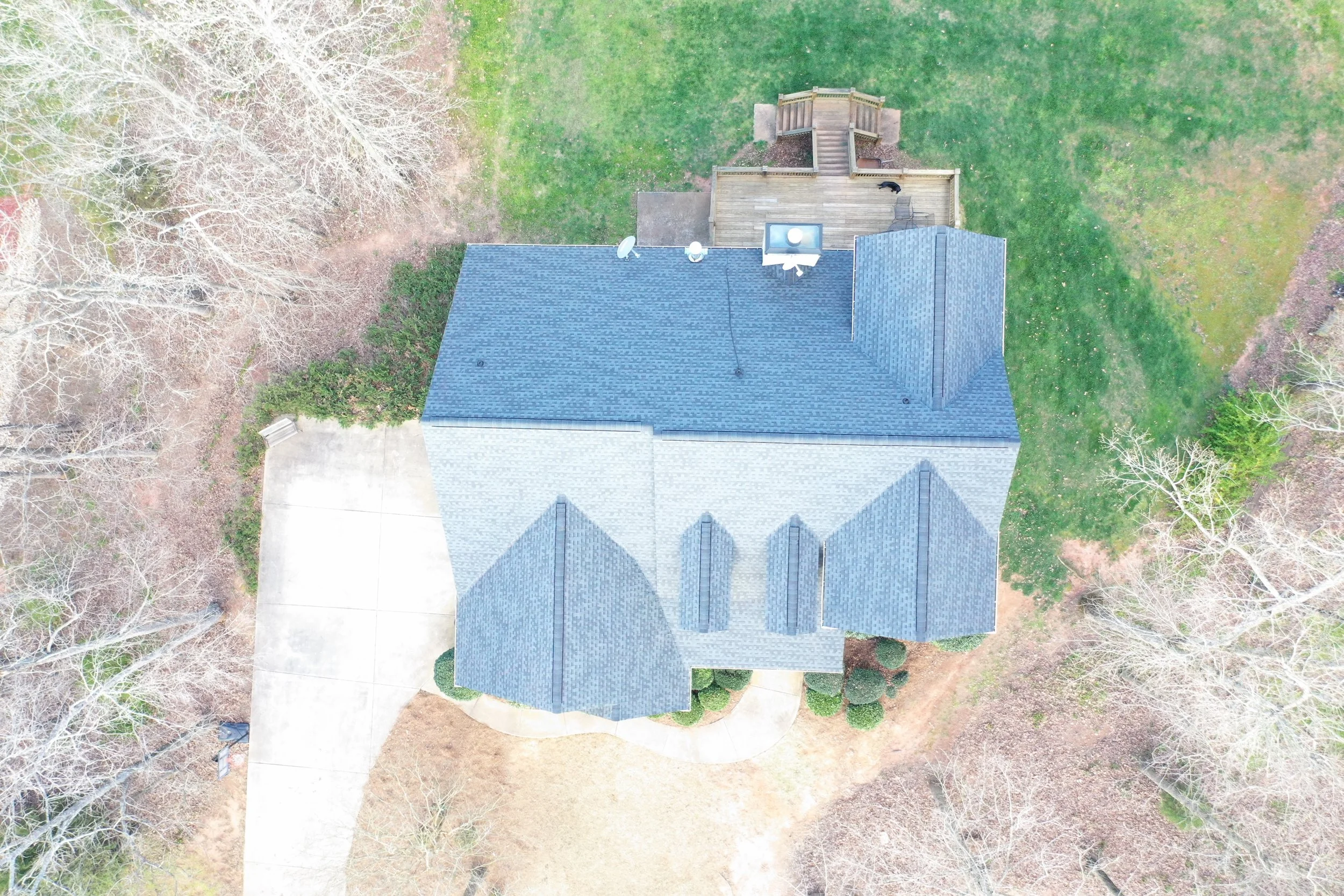 An overhead aerial view of a house with a gray shingle roof, surrounded by trees and a backyard with a wooden deck, staircase, and outdoor furniture.