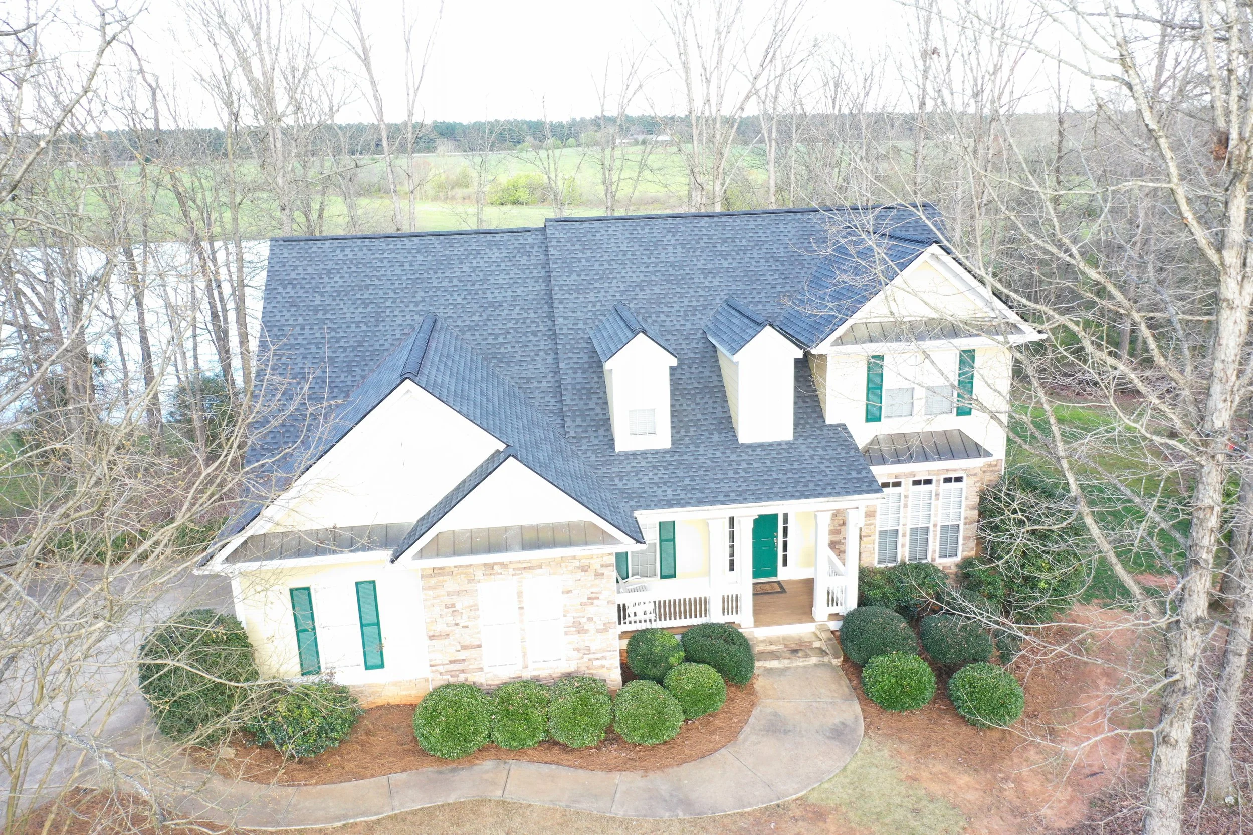 A view of a house with a dark blue shingled roof, green shutters, and a front porch, surrounded by leafless trees and neatly trimmed bushes.