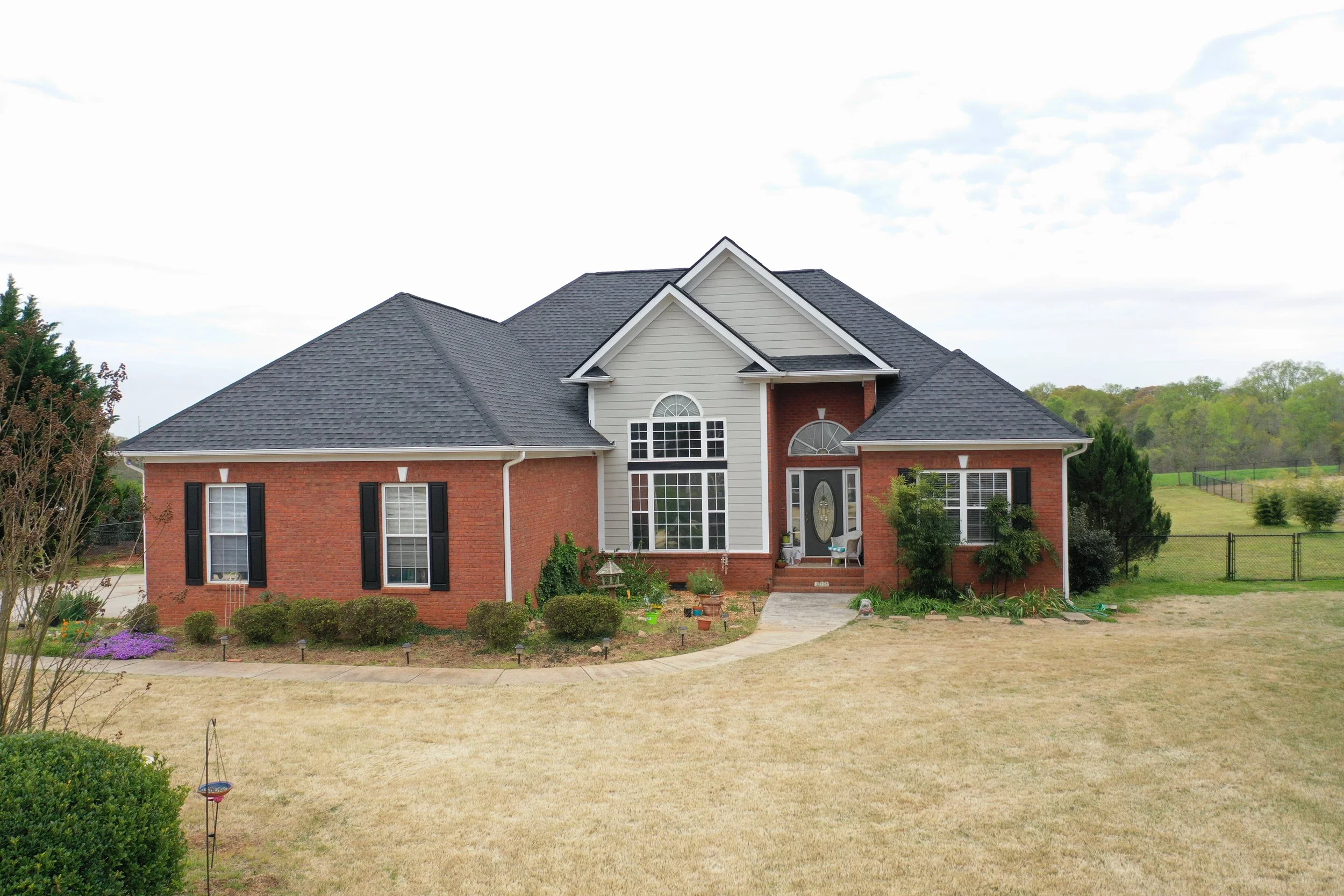 A two-story brick house with a gray shingle roof and white siding on the upper part. The house has large windows, black shutters, and a front door with a decorative glass panel. There is a small front porch with chairs and potted plants. The front ya