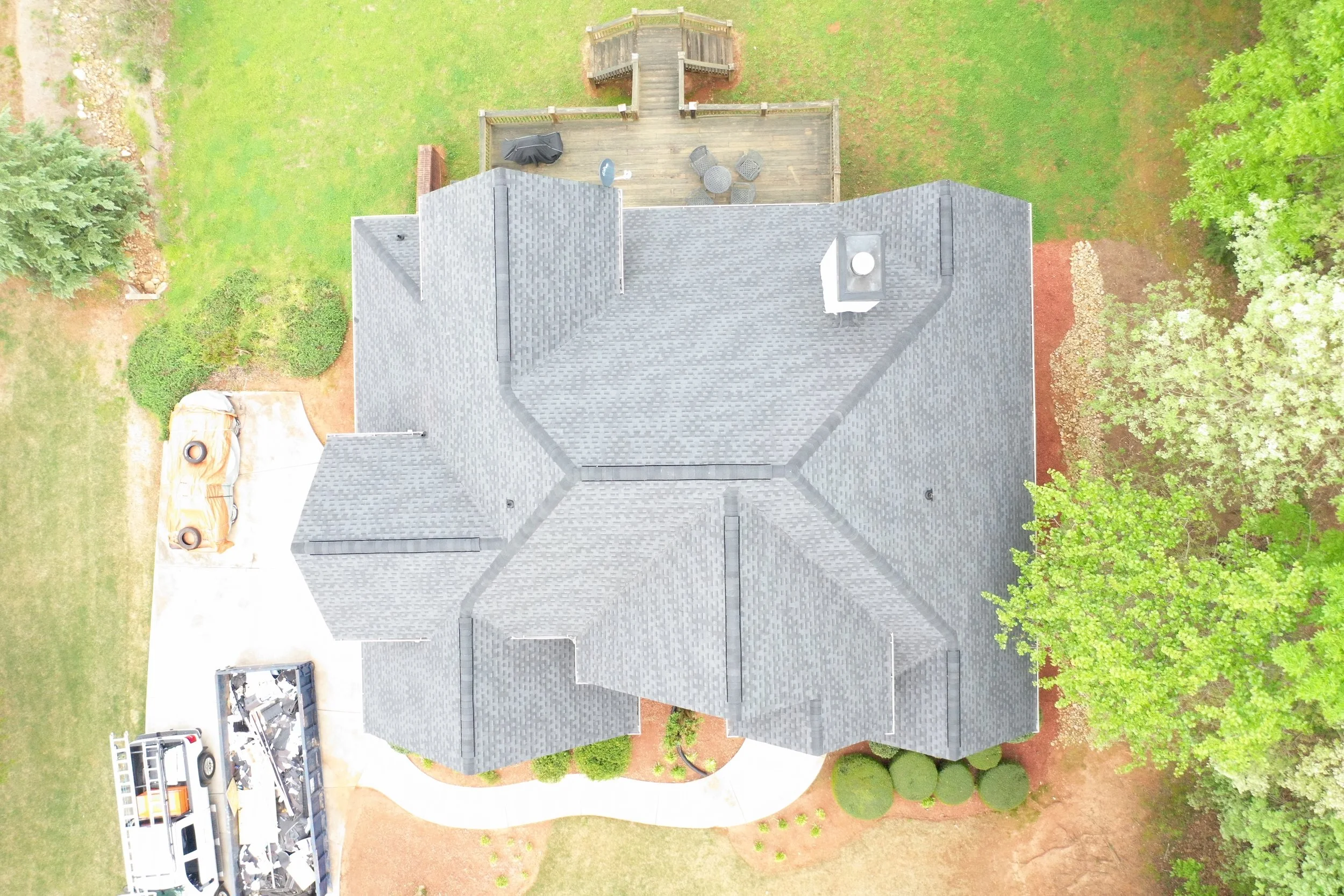 An aerial view of a house with a gray shingled roof, a wooden deck with outdoor furniture, surrounded by green grass and trees.