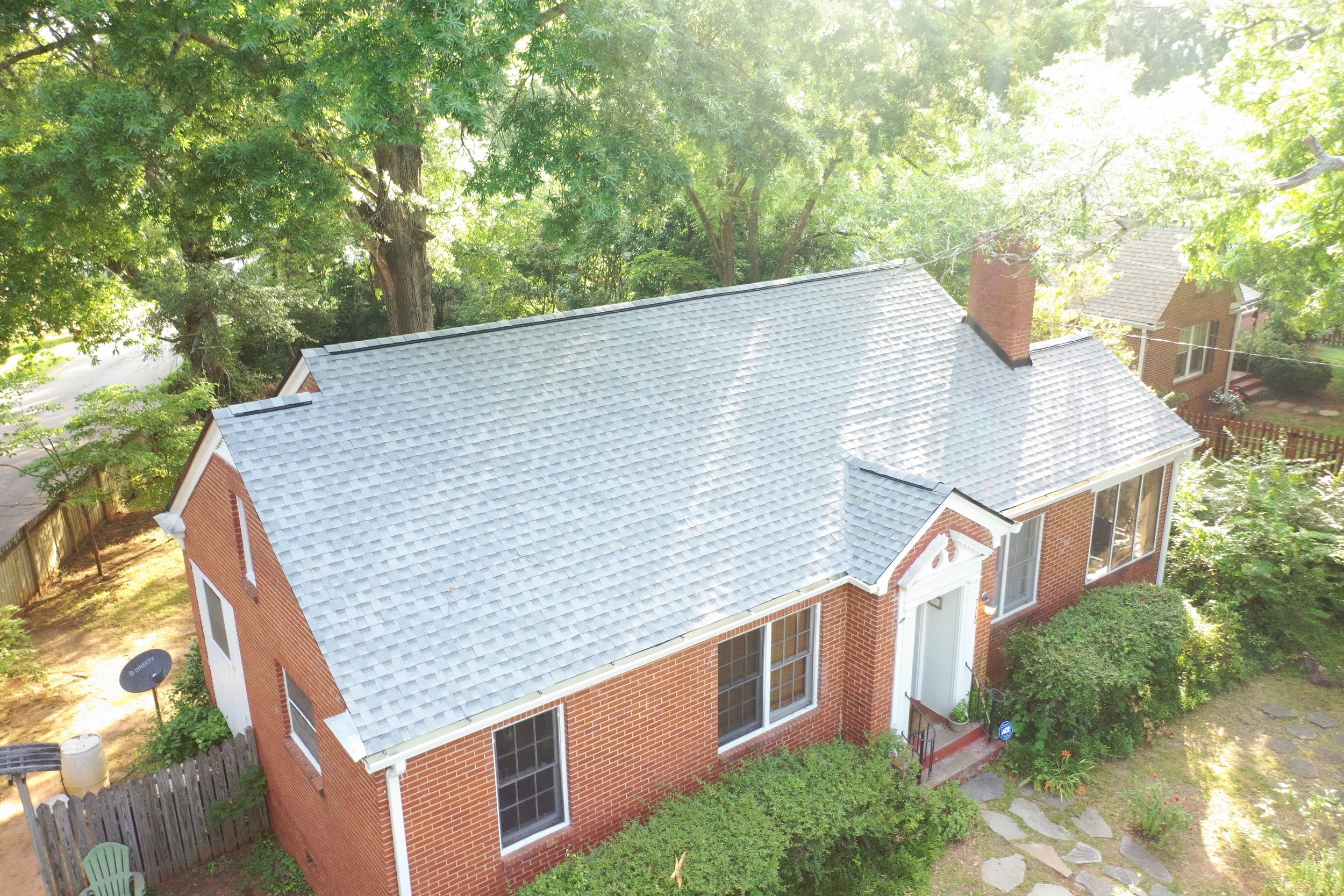 Aerial view of a brick house with a gray shingled roof, surrounded by tall green trees in a backyard with stepping stones and garden plants.