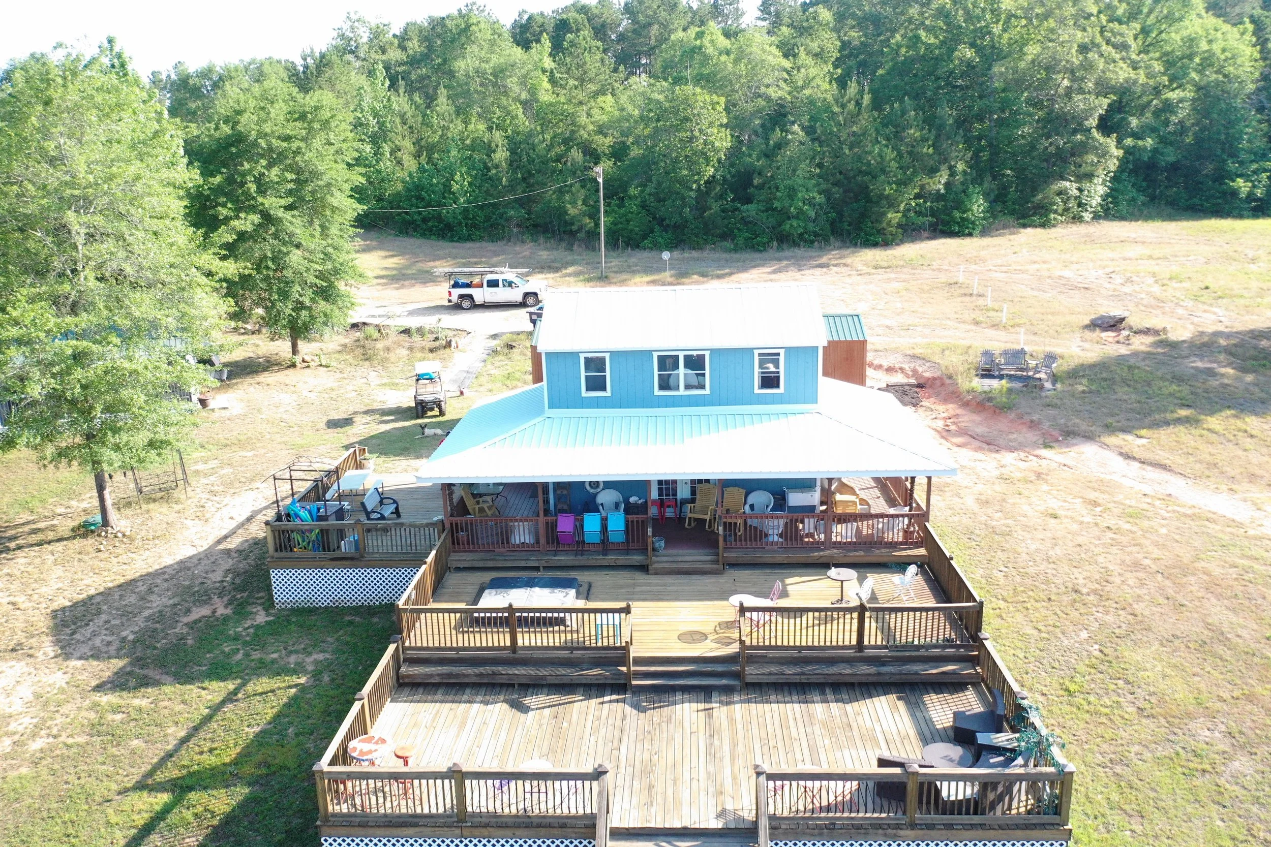 Aerial view of a backyard with a wooden deck, a small blue house with white trim, and green trees in the background.