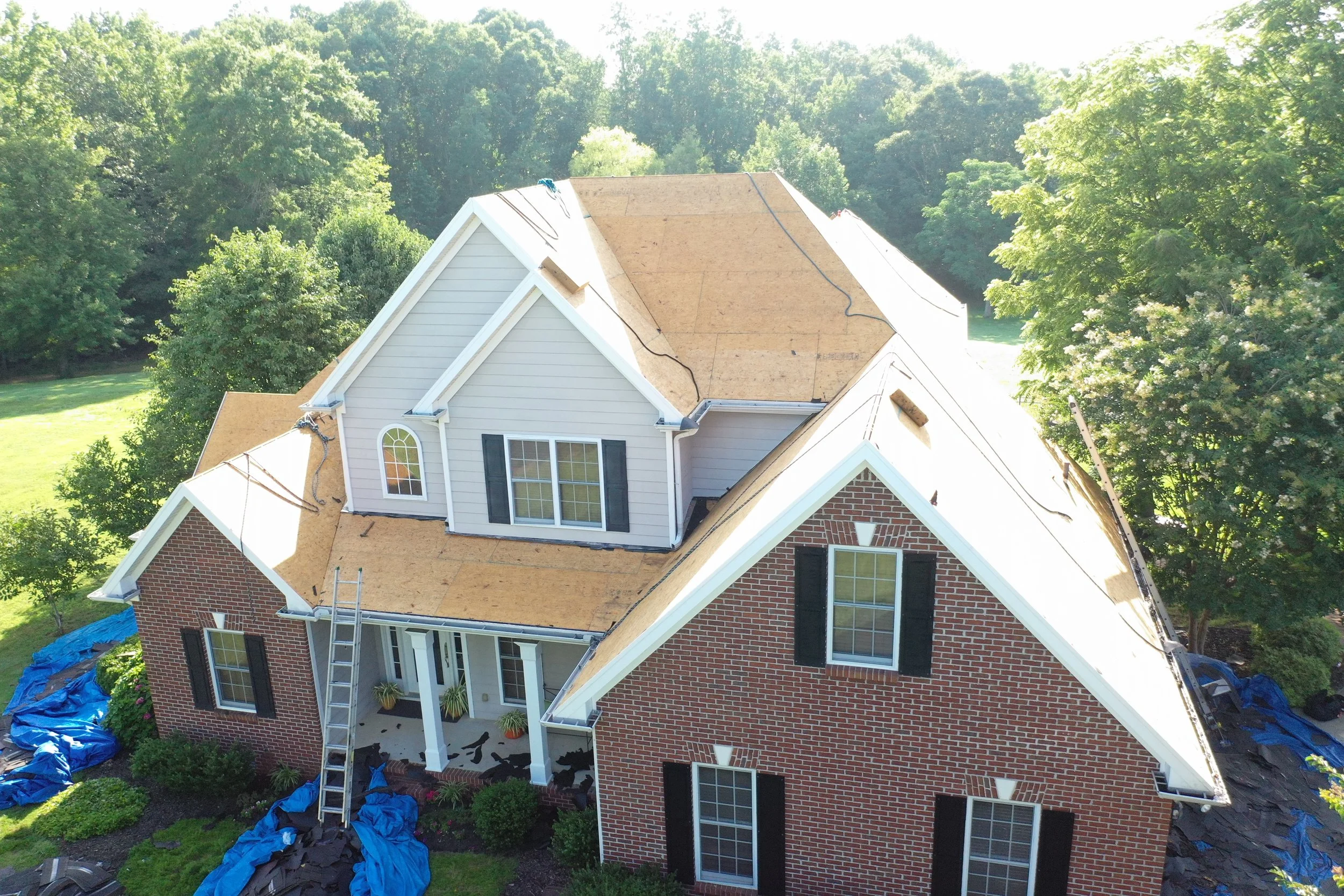 A house with a red brick exterior and black shutters undergoing roof replacement, with plywood sheathing visible on the roof and blue tarps on the ground surrounding the house.