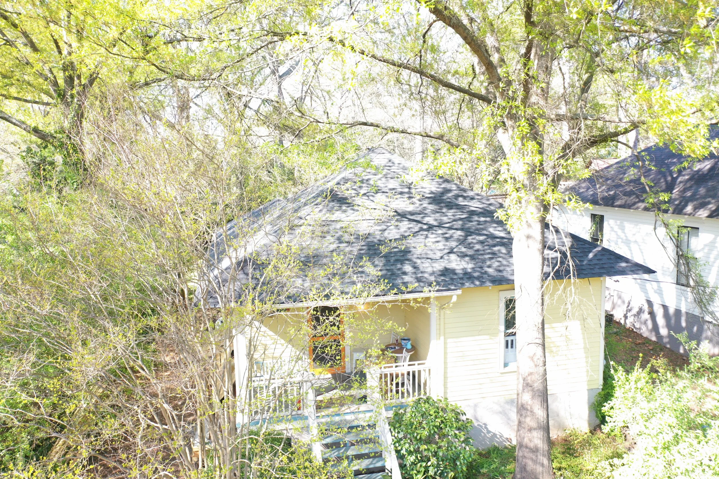 A residential house with a grey shingle roof, partially obscured by trees with green leaves, and a yellow exterior wall with a white balcony railing and staircase.