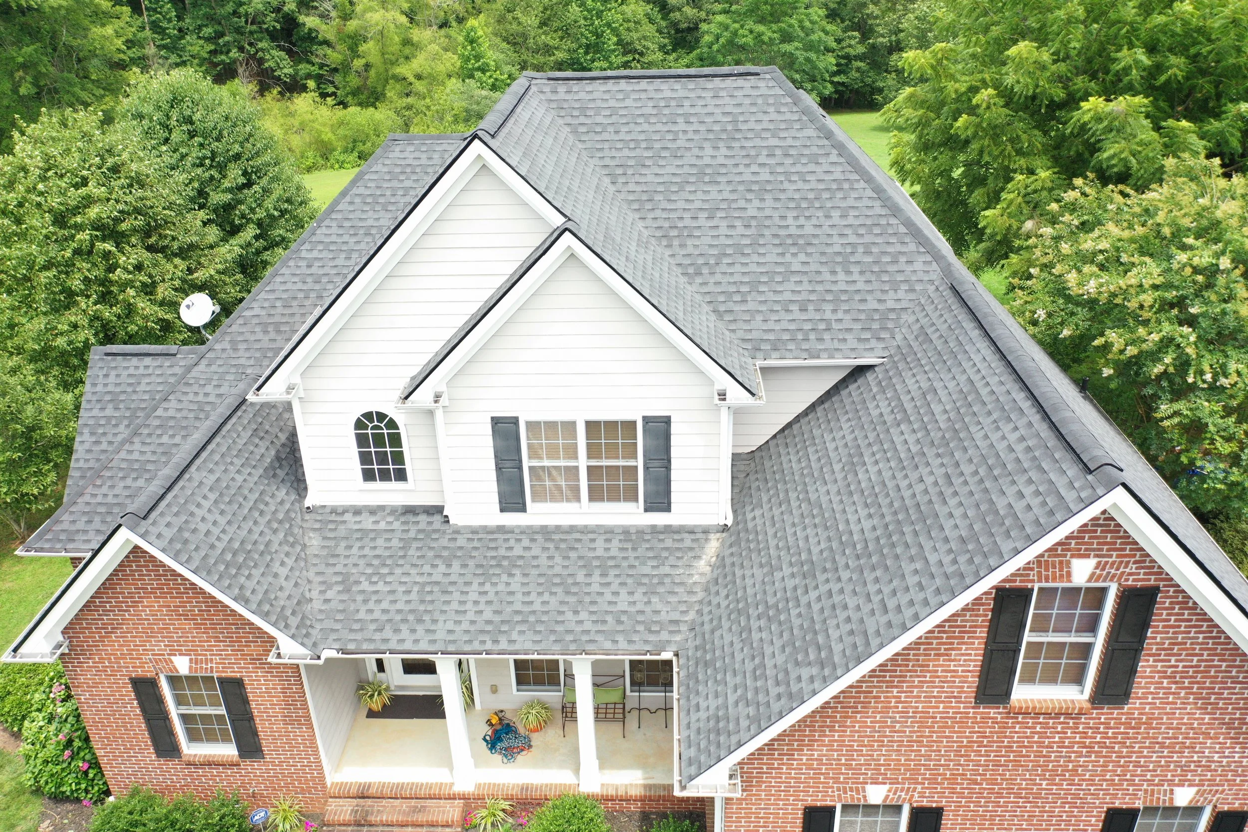 Aerial view of a house roofed by Alumni Roofing and Gutters with a grey shingle roof, white siding, brick walls, and black shutters, surrounded by green trees and a small front porch with chairs and plants.