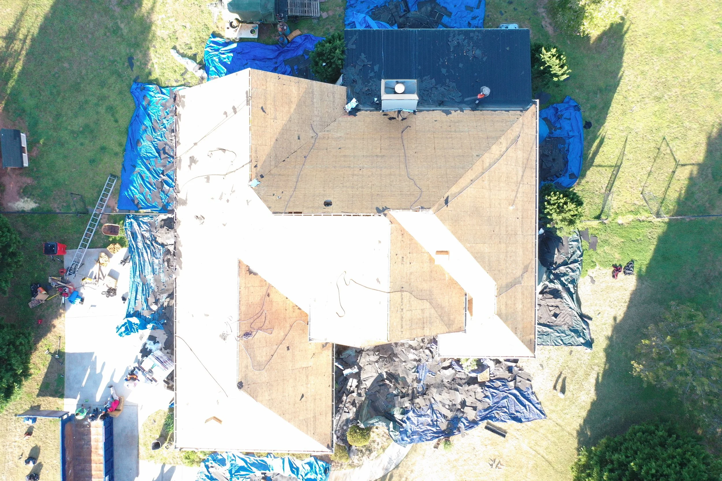 An aerial view of a house undergoing roof renovation, with sections of old roofing removed and new roofing being installed. Blue tarps cover parts of the yard and roof, and construction tools and materials are scattered around.