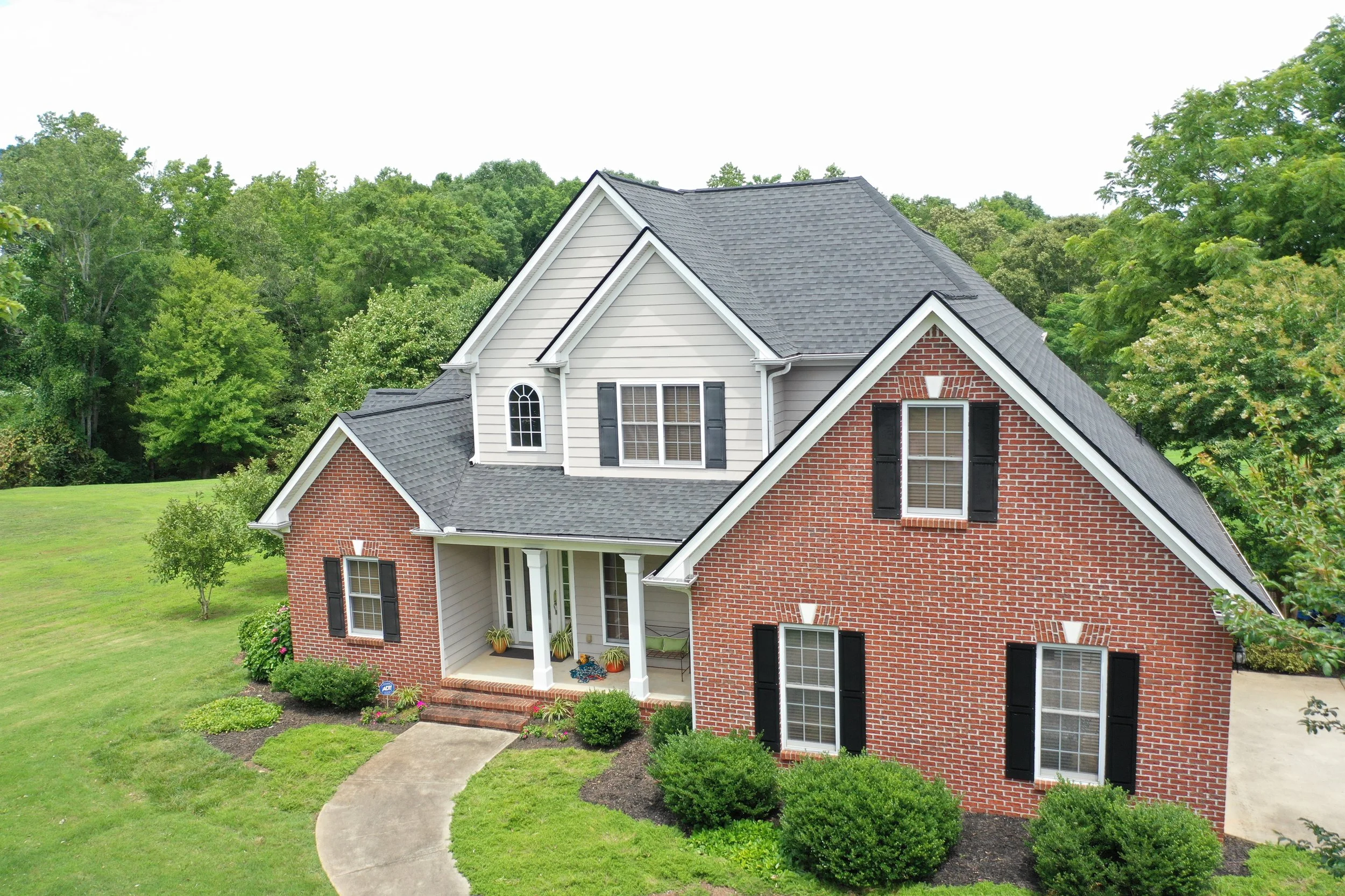 A two-story house with a brick and siding exterior, black shutters, a front porch with potted plants, surrounded by a well-maintained lawn and trees, under a cloudy sky.