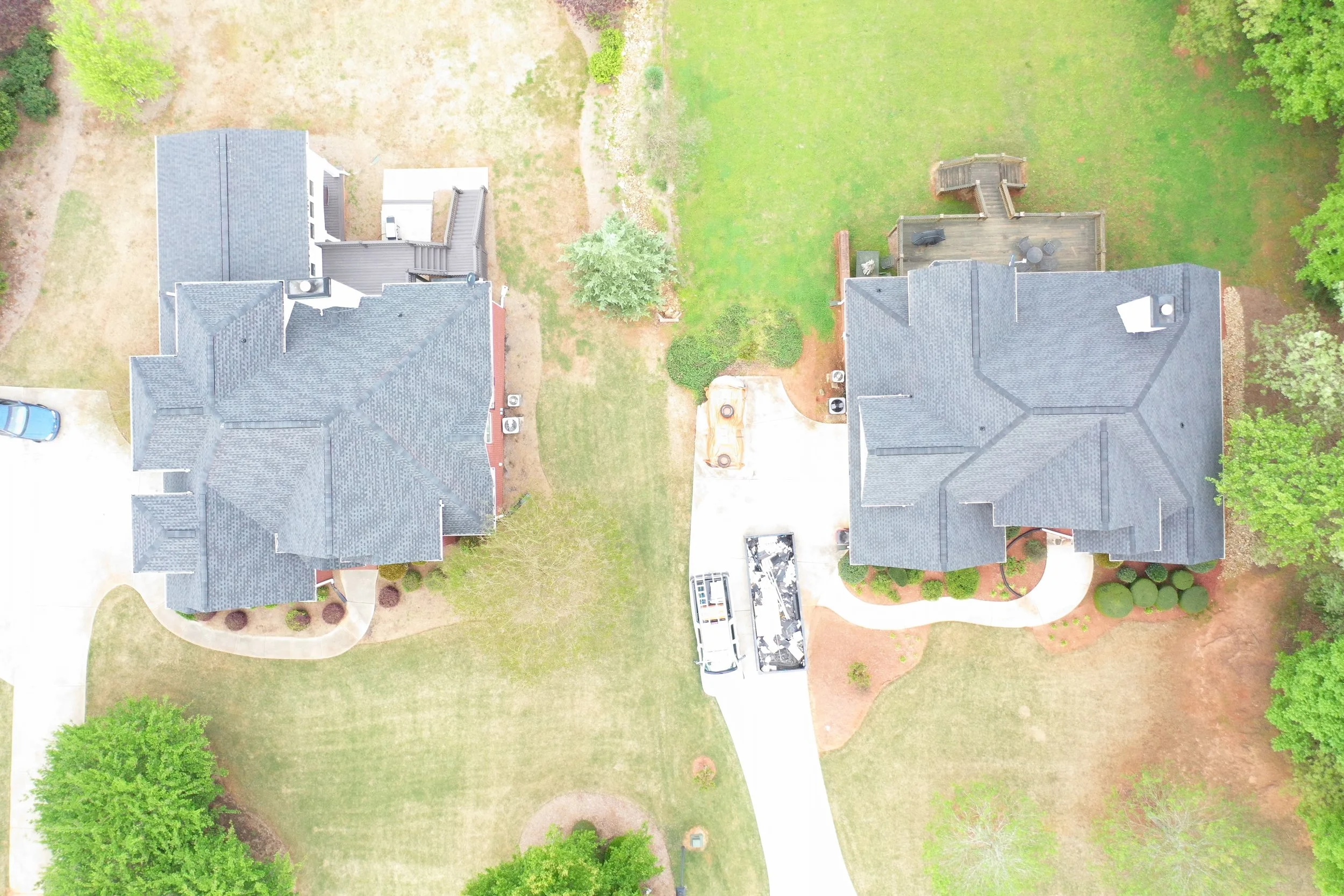 An aerial view of a suburban neighborhood showing two houses, a driveway with a parked car, a wooden deck, and green lawns with trees.