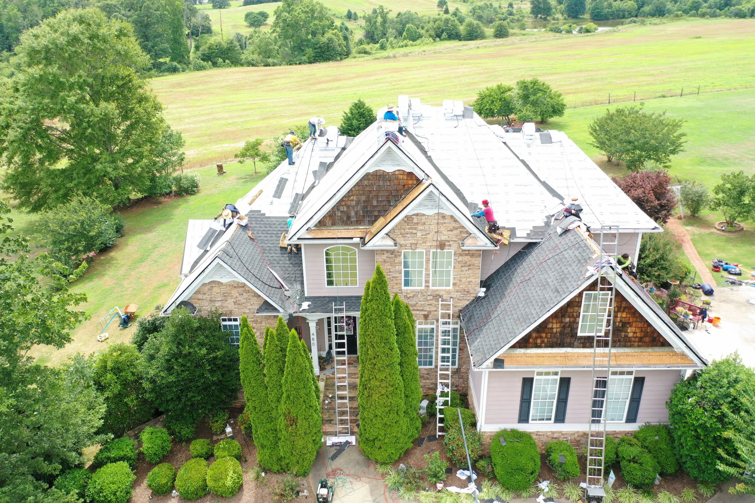 Overhead view of a house with a roof undergoing repair, with multiple workers on the roof, ladders, and construction tools, surrounded by green trees and a large grassy field in the background.