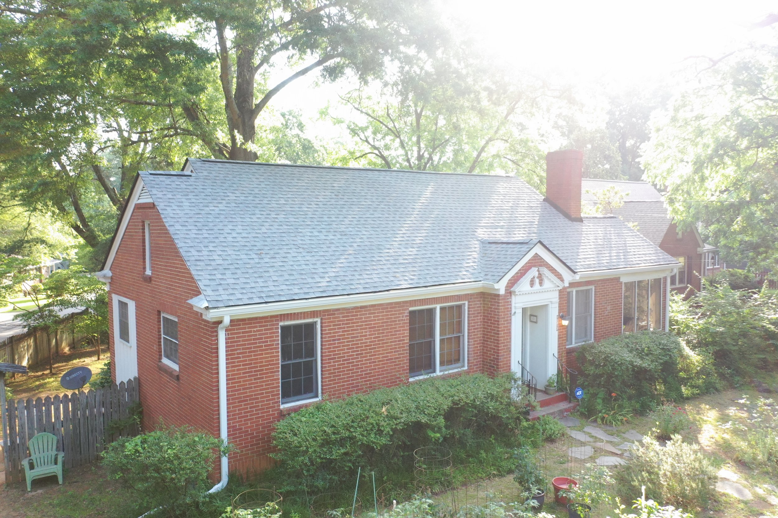 A two-story brick house with large windows, a blue shingle roof, and a chimney. The backyard has a small garden, a pathway, and trees surrounding the house. The sun is shining brightly, creating a glowing effect.
