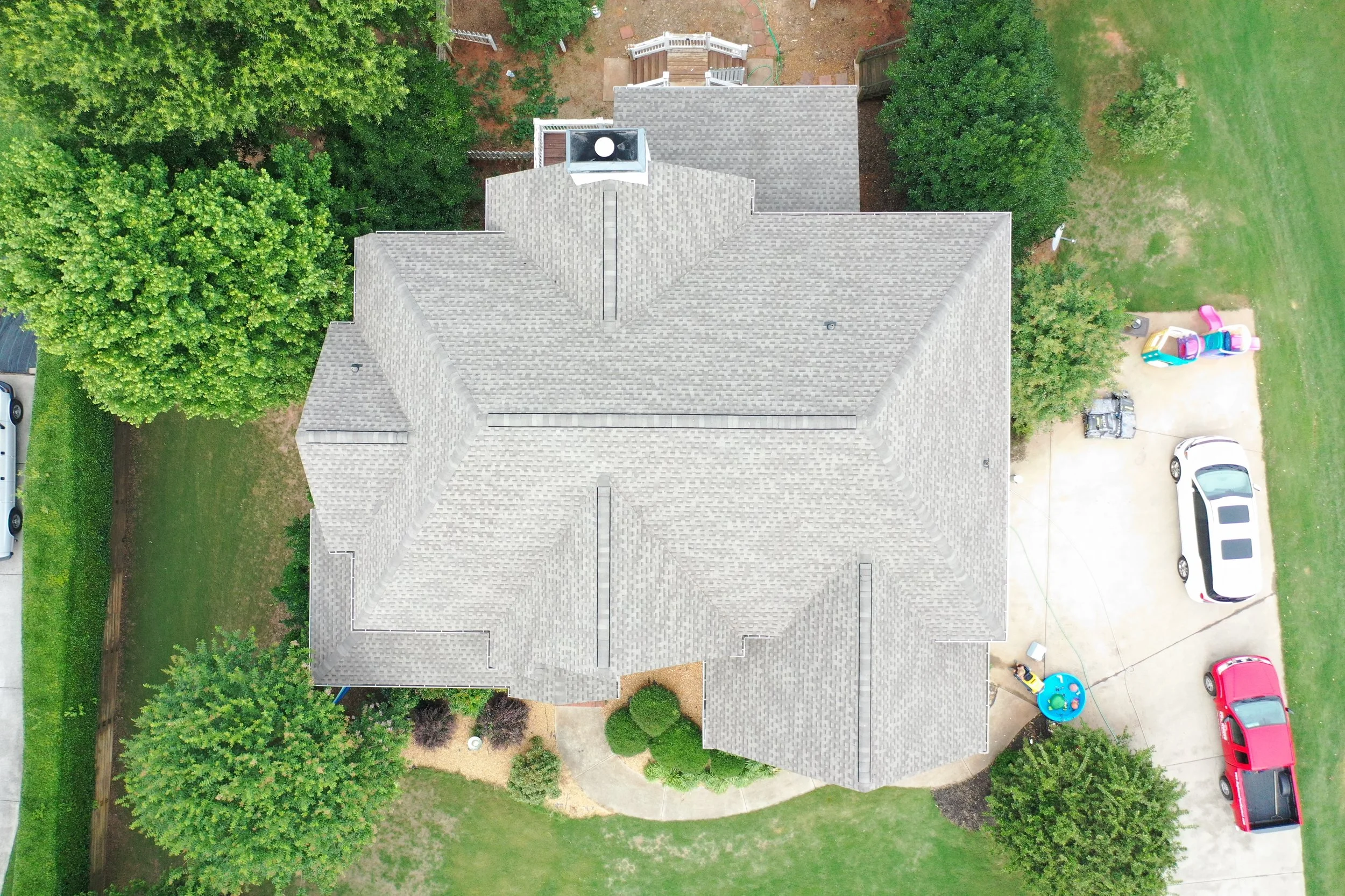 Aerial view of a house with a gray shingle roof, surrounded by green trees and a backyard with a wooden deck. The driveway has several parked cars and children's toys.