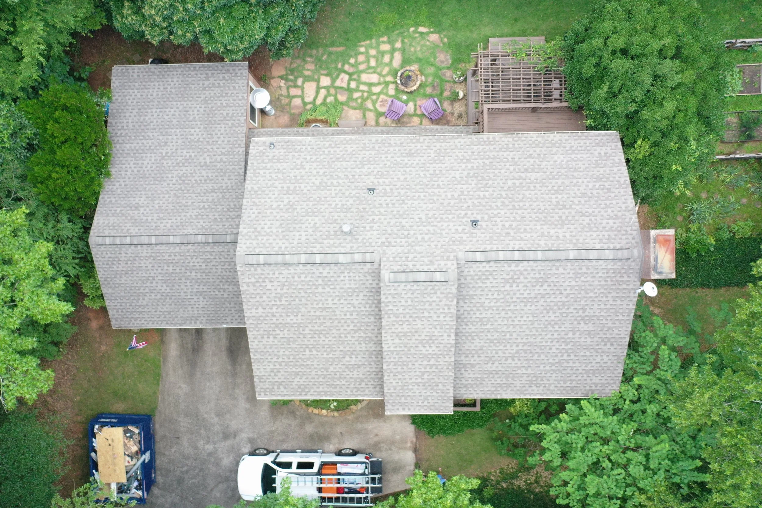 An aerial view of a backyard showing a house with a gray shingled roof, a driveway, two parked vehicles, a blue trailer, and a patio area with two purple chairs, a fire pit, and surrounding greenery.