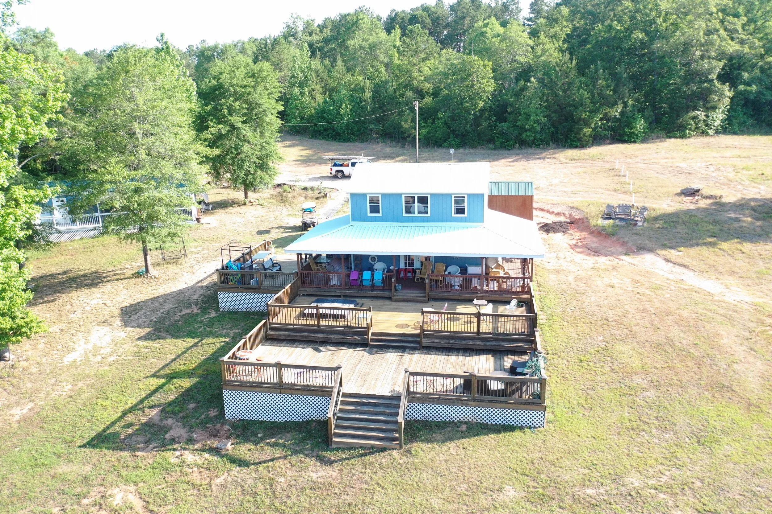 Aerial view of a house with a deck in a grassy yard, surrounded by trees and open fields, with a pickup truck parked nearby.