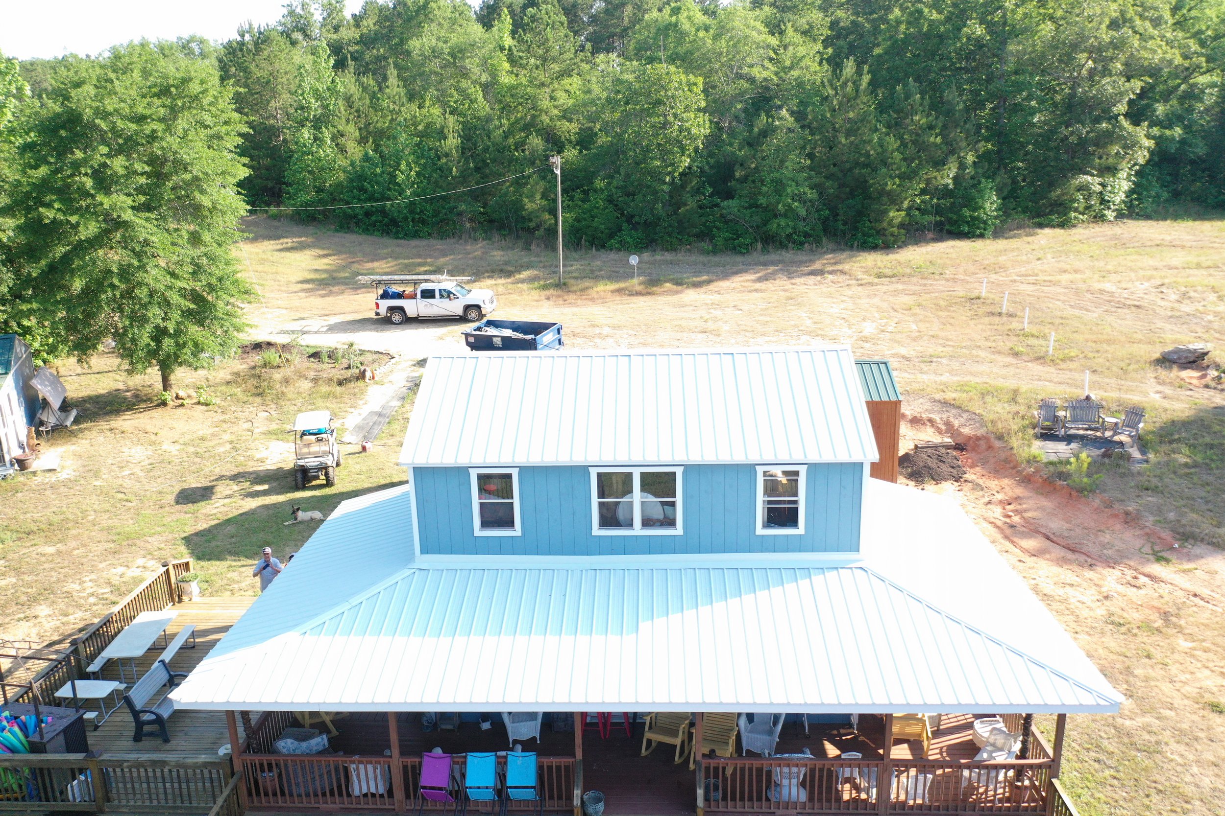 Aerial view of a backyard with a blue house, deck with chairs, and a grassy area with trees and shrubs in the background.