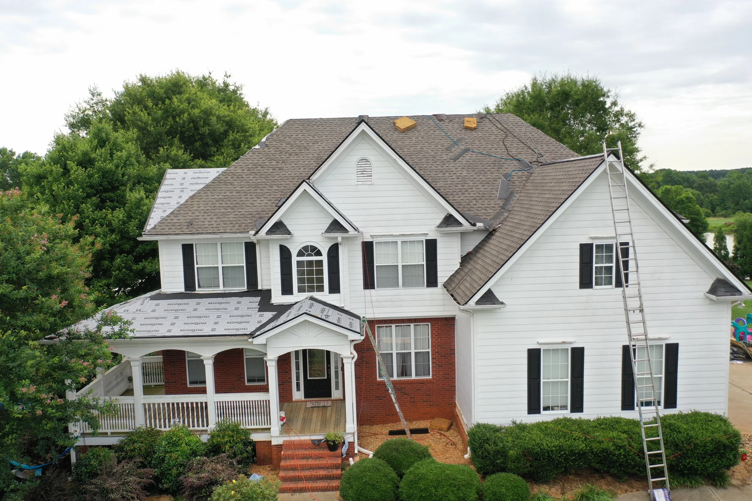 Front of a two-story house with white siding, black shutters, and a brick foundation. The roof is being replaced or repaired, with some shingles missing and new sheathing visible. A ladder leans against the right side of the house, and there is a small porch with a white railing and steps leading to the front door. Surrounding the house are trees, shrubs, and a landscaped yard.
