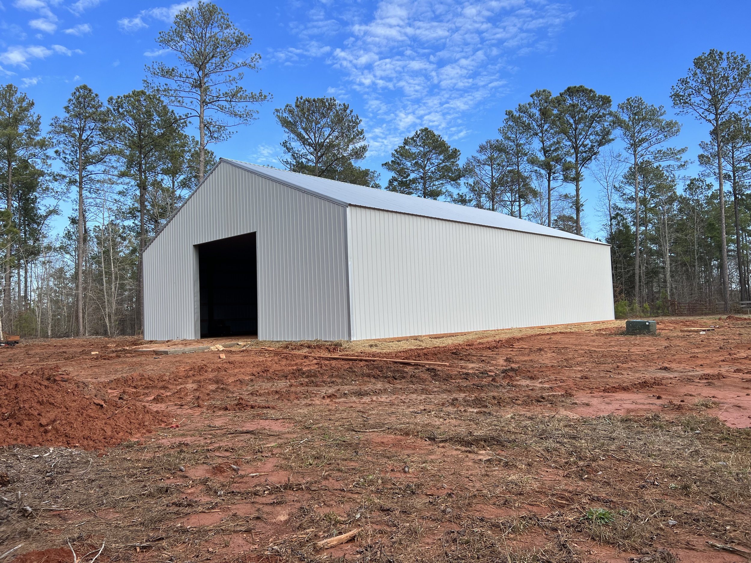 A white metal building with an open entrance, set against a backdrop of tall pine trees under a blue sky with scattered clouds. The ground is reddish-brown, uneven soil with patches of grass.