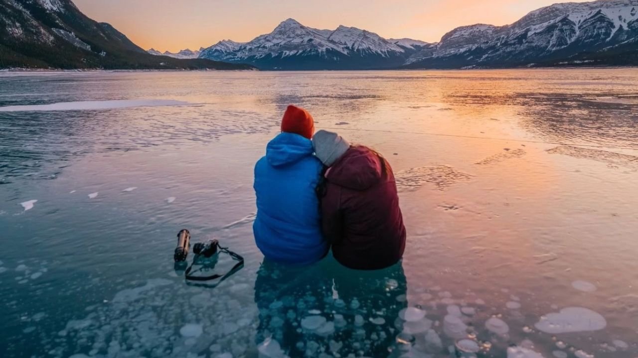 Visitors sitting on Abraham Lake ice bubbles during winter sunset with Canadian Rockies mountains in the background.