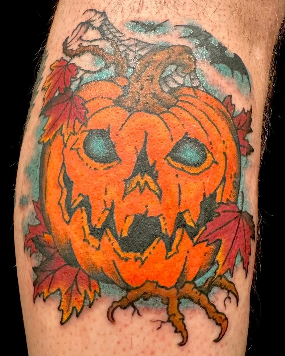 Color tattoo of a pumpkin with bats and a moon on a lower leg.