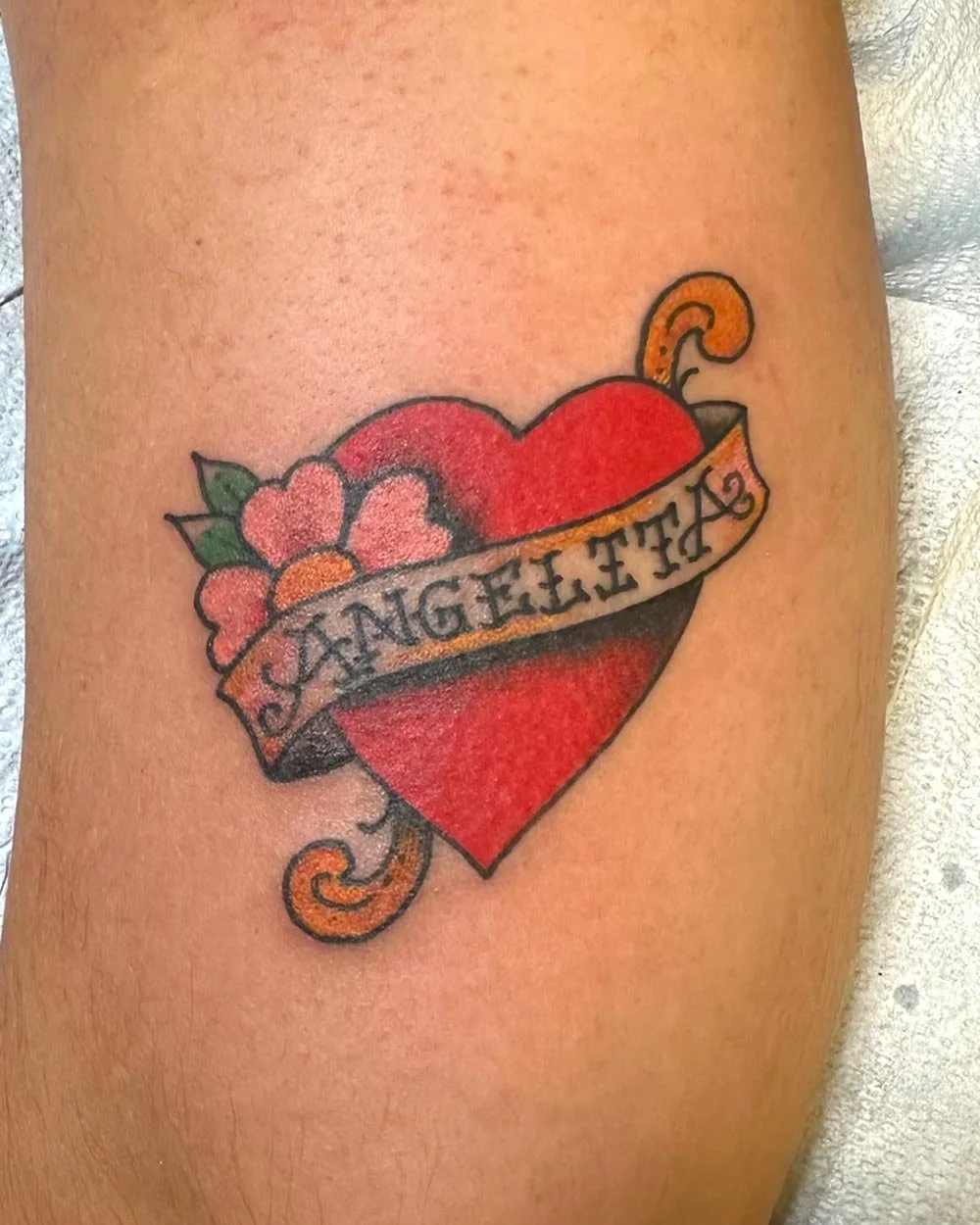 Traditional color tattoo of a heard and a banner with a small pink flower on the back of an arm.