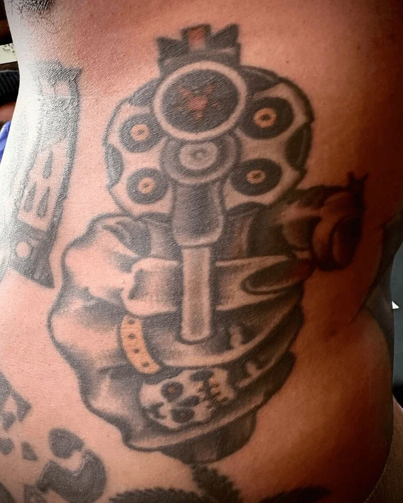 Rib tattoo in color by John Reardon of a hand with rings holding a gun.