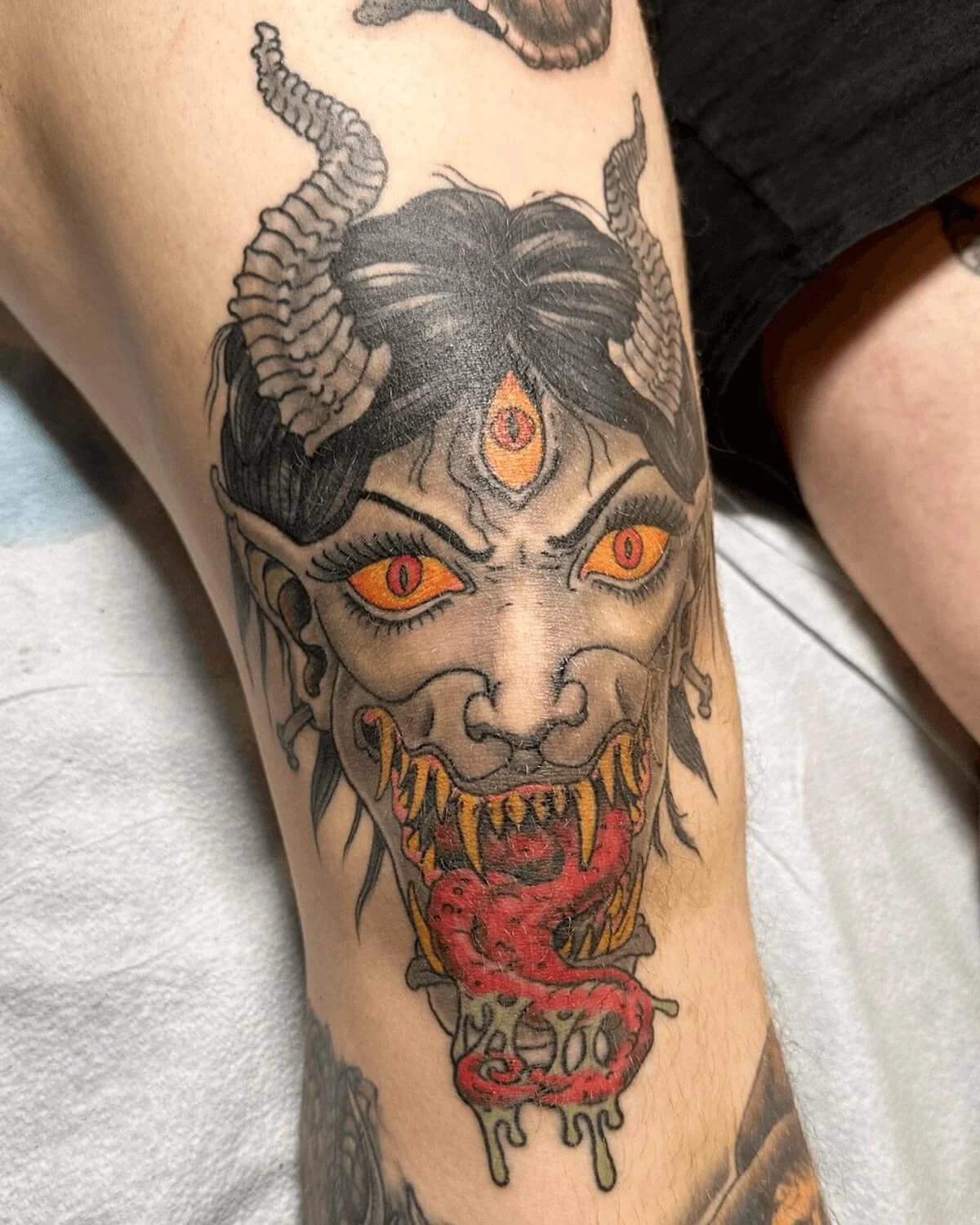 Color knee tattoo of a demon with three eyes, horns, and long tongues.