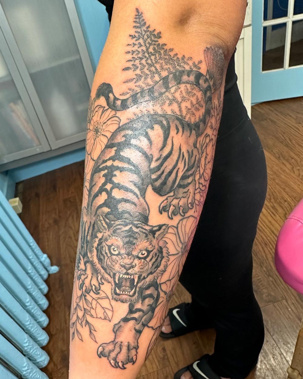 Tiger and fauna tattoo on forearm in black and grey in illustrative realistic styles.